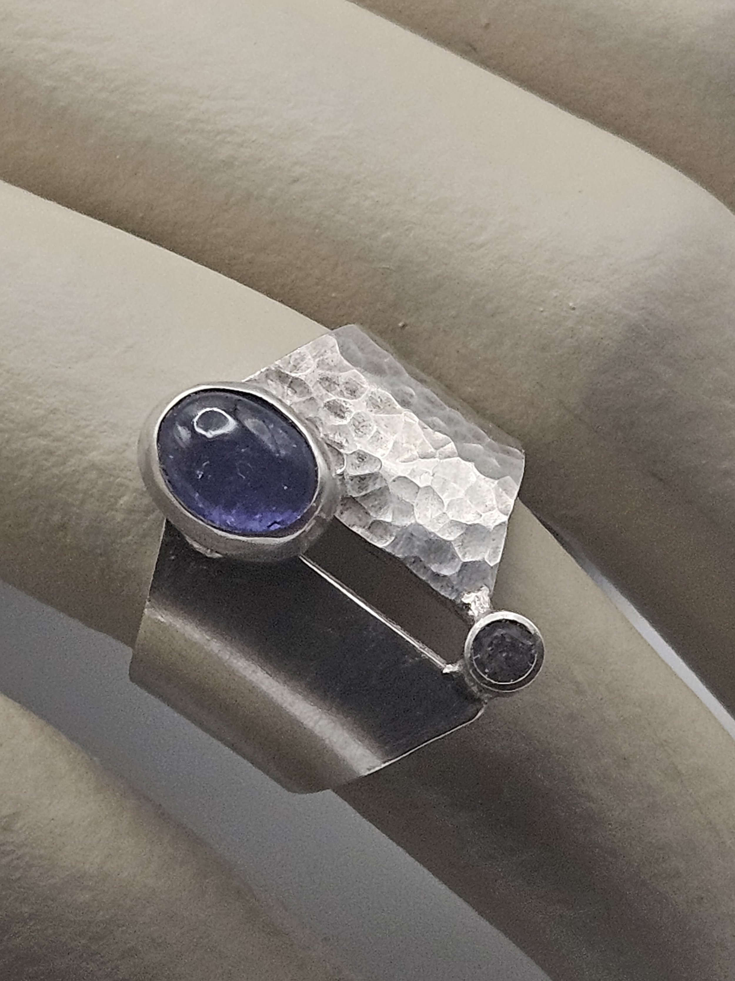 Close-up of a silver hand-crafted ring with hammered texture, featuring two oval-shaped dark blue stones, one larger and one smaller, set in silver bezels, resting on a neutral surface.