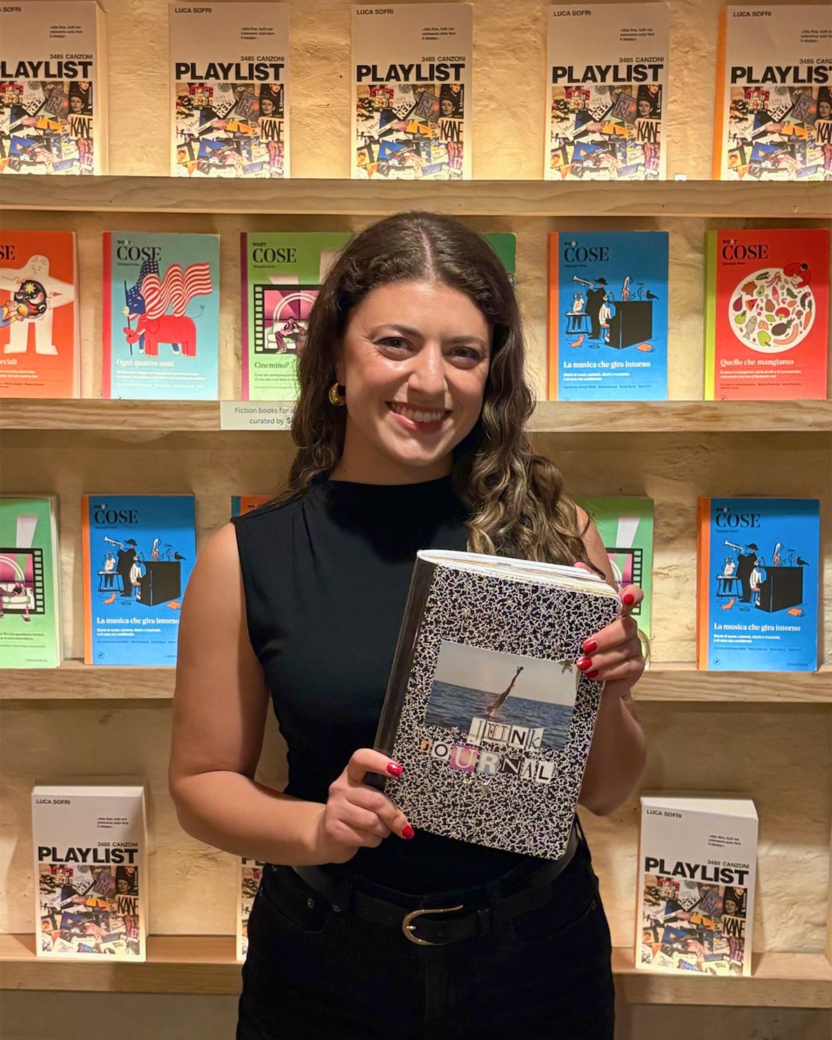 Collageria founder, Francesca Maida, holds her junk journal and smiles in front of a colorful wall of books. Her hair is down, she is wearing all black, and smiling.