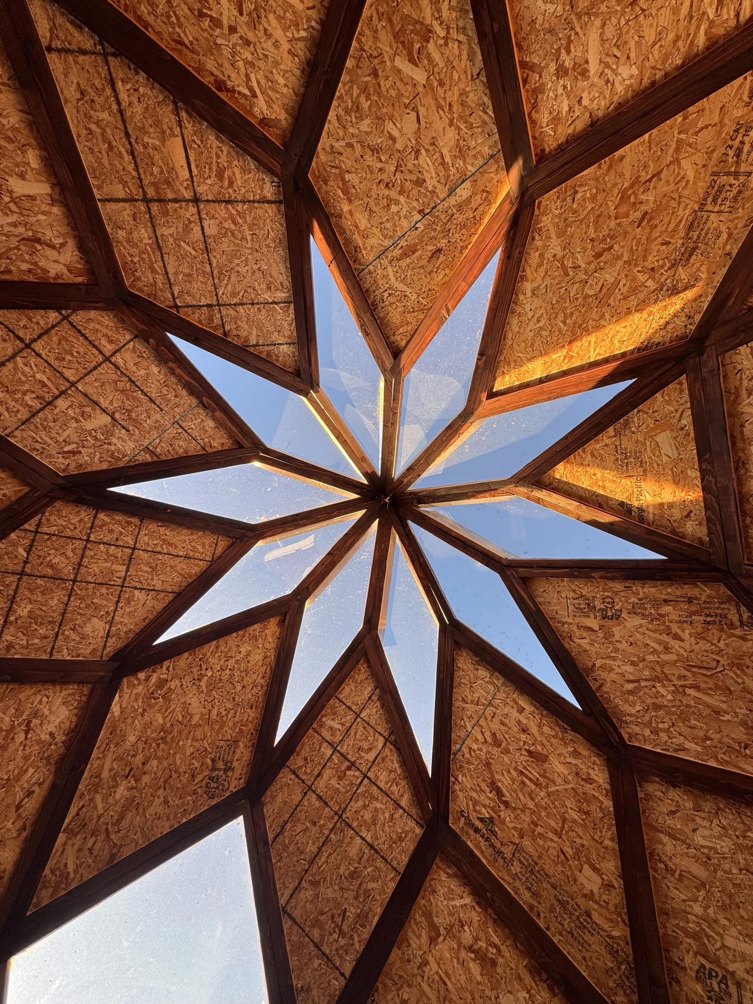 View of a star-shaped skylight made of oriented strand board and glass panels from inside a wooden structure, looking up at a clear blue sky.