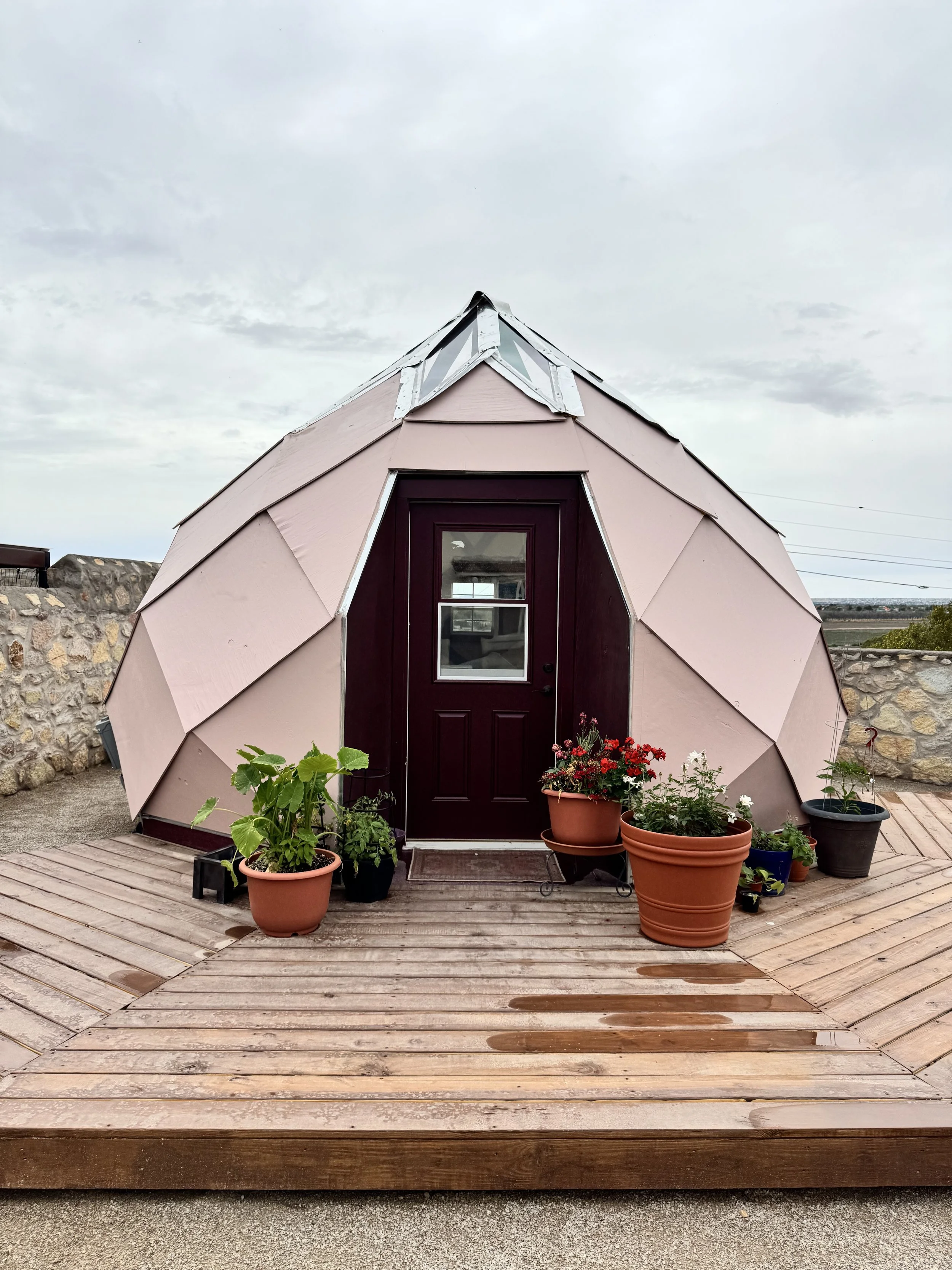 A small geodesic dome house with a mauve exterior, a dark maroon door, and potted plants outside on a wooden deck, under a cloudy sky.