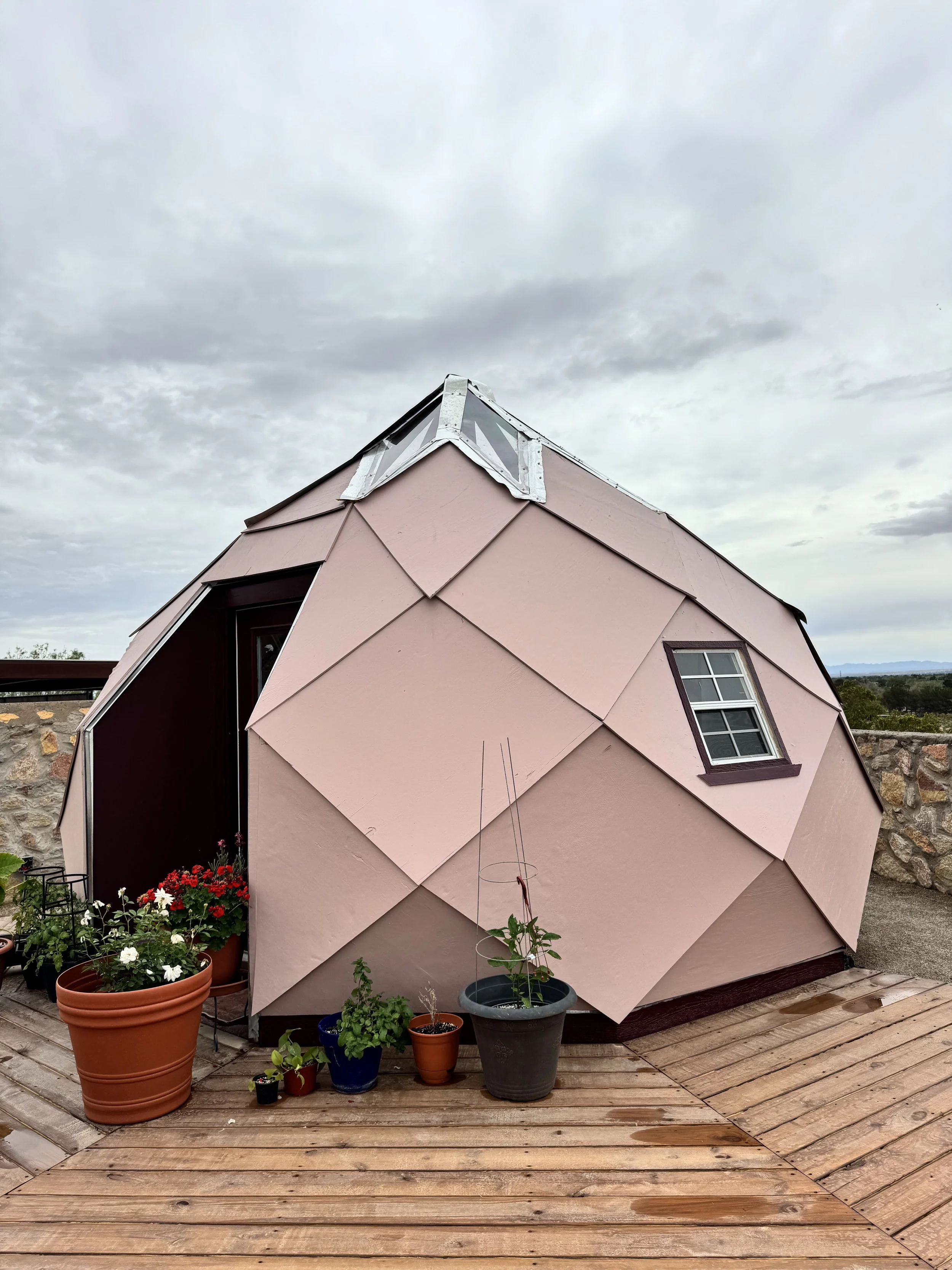 Small, pink, dome-shaped house with a geometric design, small window, and plants in pots gathered outside, set on a wooden deck under a cloudy sky.