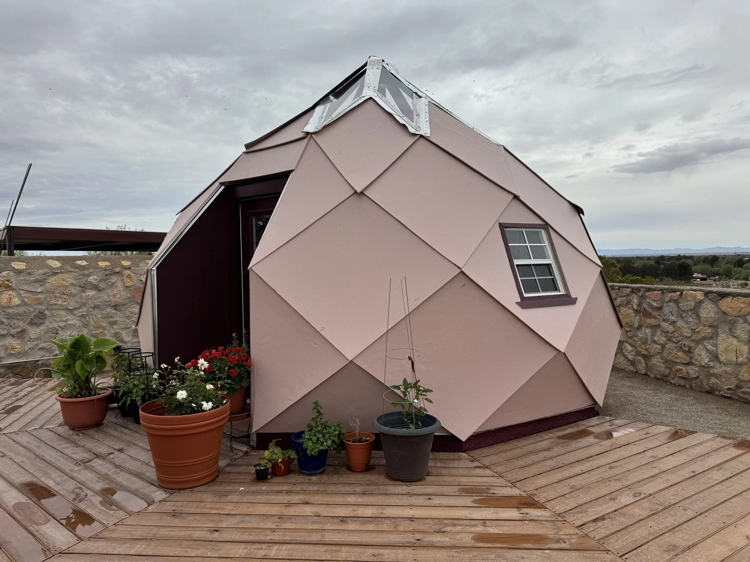 A geodesic dome-shaped tiny house with pink exterior panels and small windows, situated on a wooden deck with potted plants and flowers, with a stone wall and cloudy sky in the background.