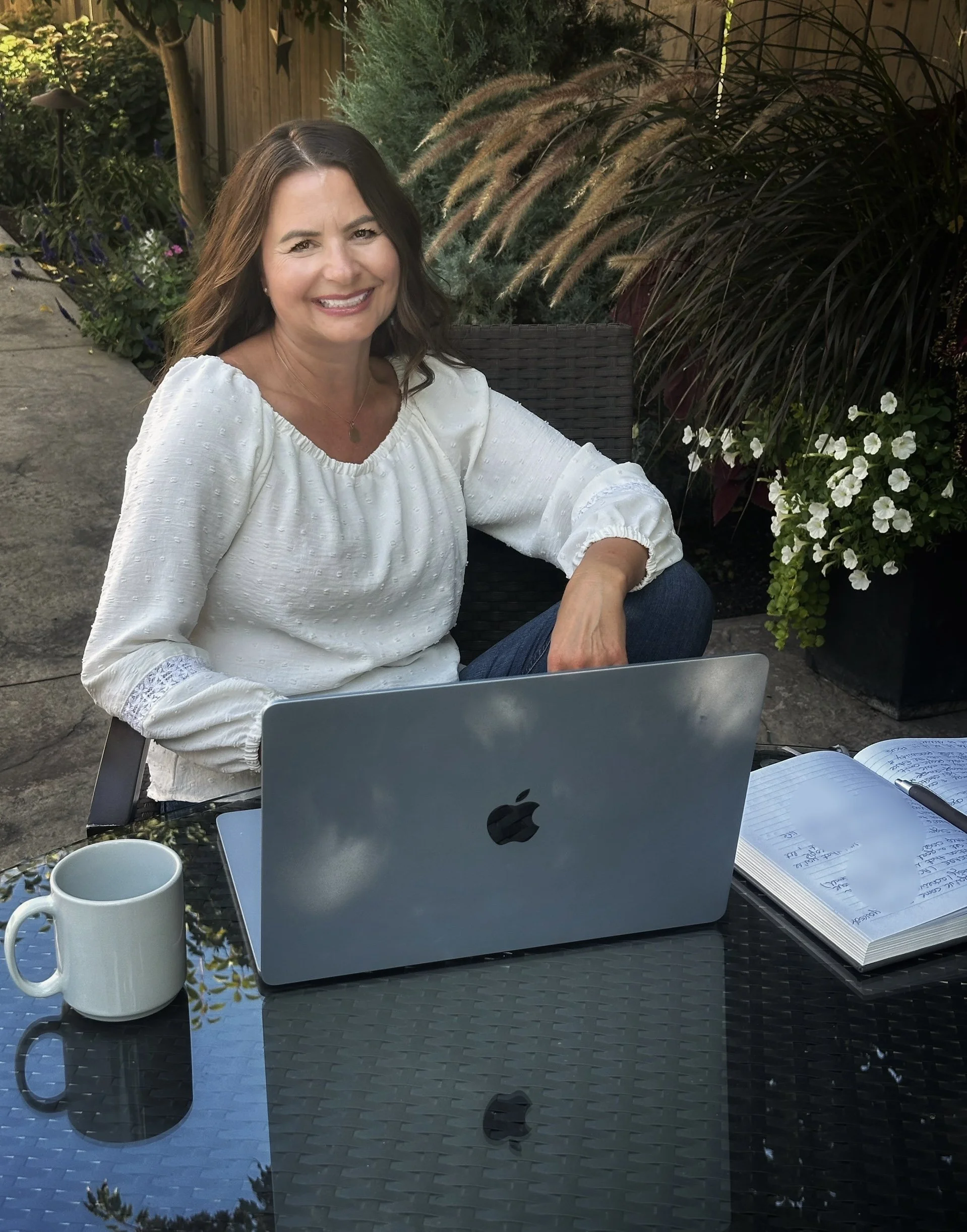 A woman sitting outdoors at a glass table with a silver MacBook, a white coffee mug, and an open notebook with handwritten notes.