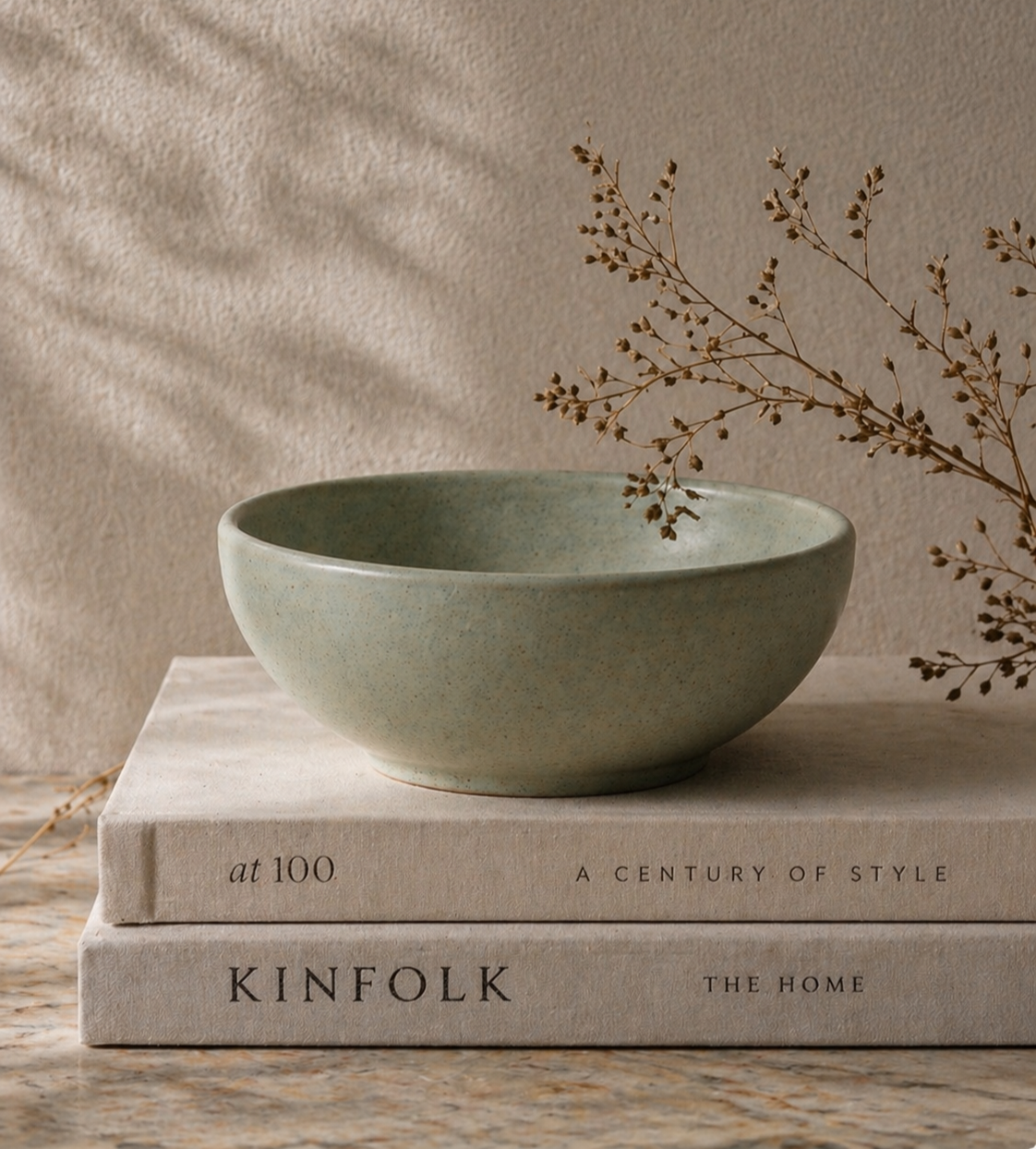 A light green ceramic bowl resting on two stacked beige books, with a dried branch of small seed pods next to it, on a marble surface with a textured beige background.