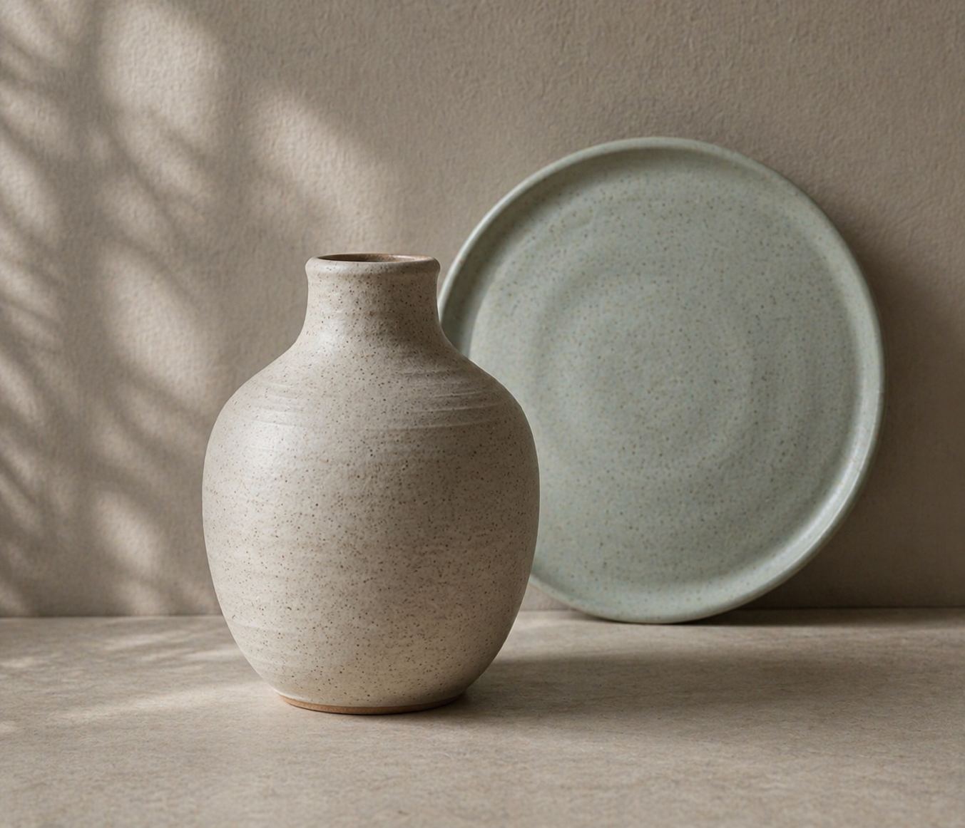 A beige speckled ceramic vase with a narrow neck and rounded body, placed on a neutral surface with a light green speckled plate leaning against the wall behind it.