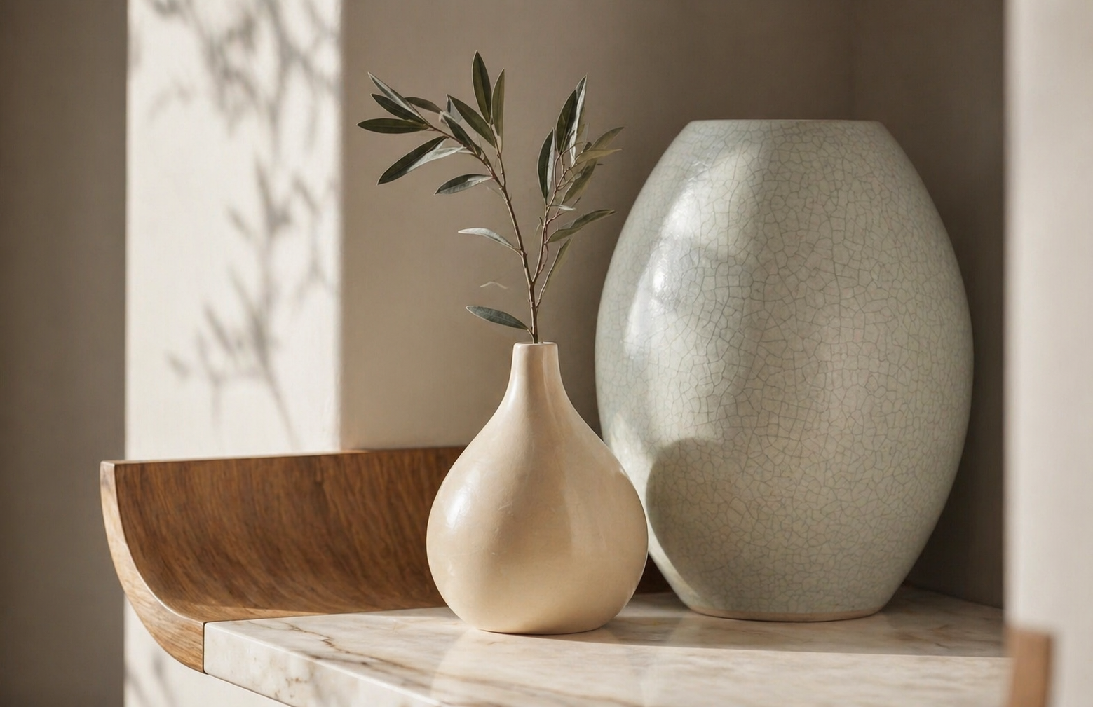 Close-up of a small beige vase with green leaves, a large crackled beige vase, and a curved wooden accessory on a marble surface, with shadows cast on a light-colored wall.