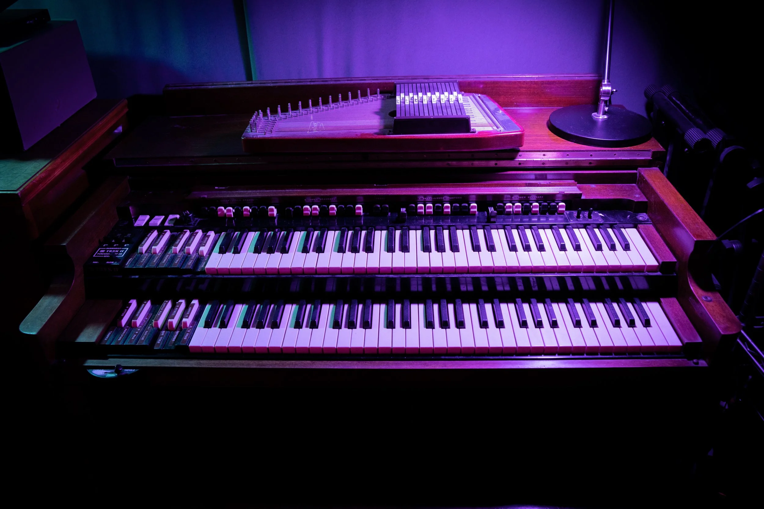 A vintage electronic organ with multiple keyboards, switches, and controls, illuminated by purple and pink stage lighting, with a small desk lamp and other equipment nearby.