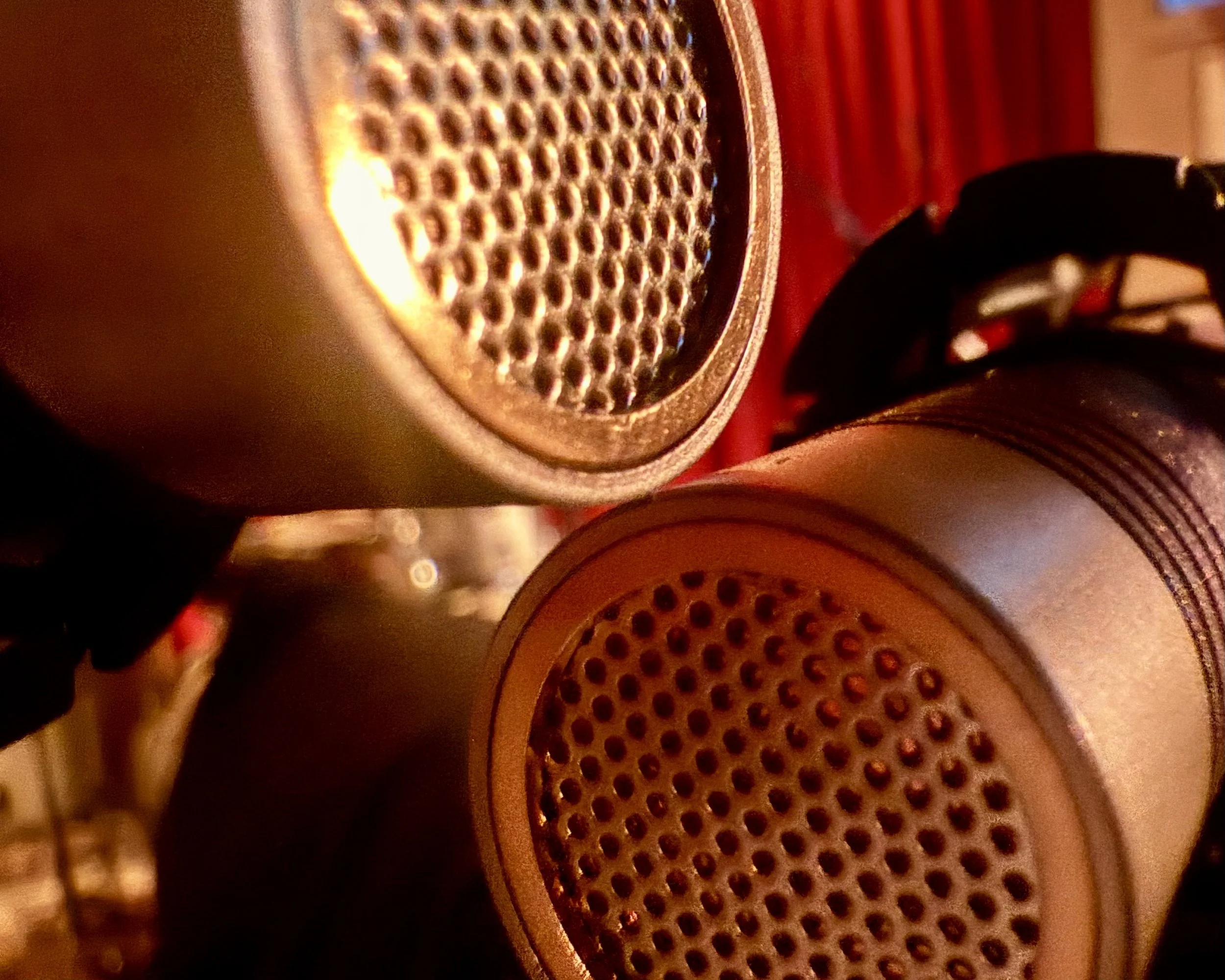 Close-up of two microphones with gold-colored grilles, one positioned in front of the other, against a background of red curtains and musical equipment.
