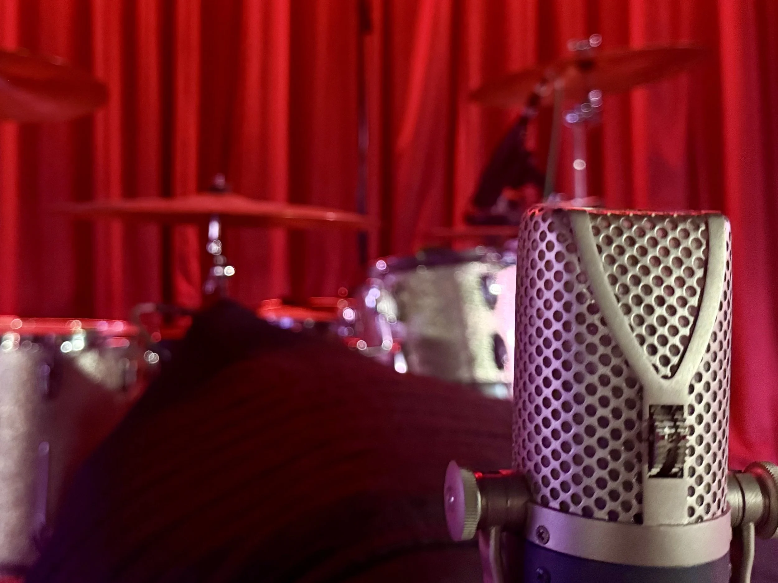 Close-up of a vintage microphone with drum set in background, red curtain backdrop.