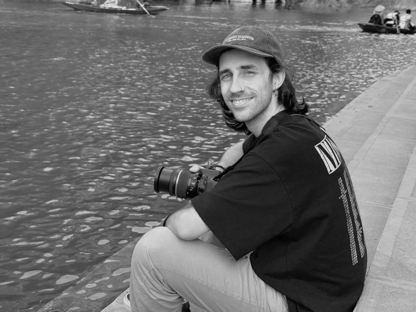 A smiling man with long hair, wearing a cap, sitting on a concrete bank by the water, holding a camera, with boats in the background.
