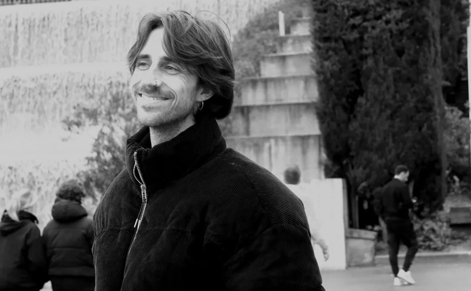 A black-and-white photo of a smiling man with medium-length hair, a goatee, earrings, and a zip-up jacket, standing outdoors with other people in the background, some walking and some seated, with trees and a wall behind him.