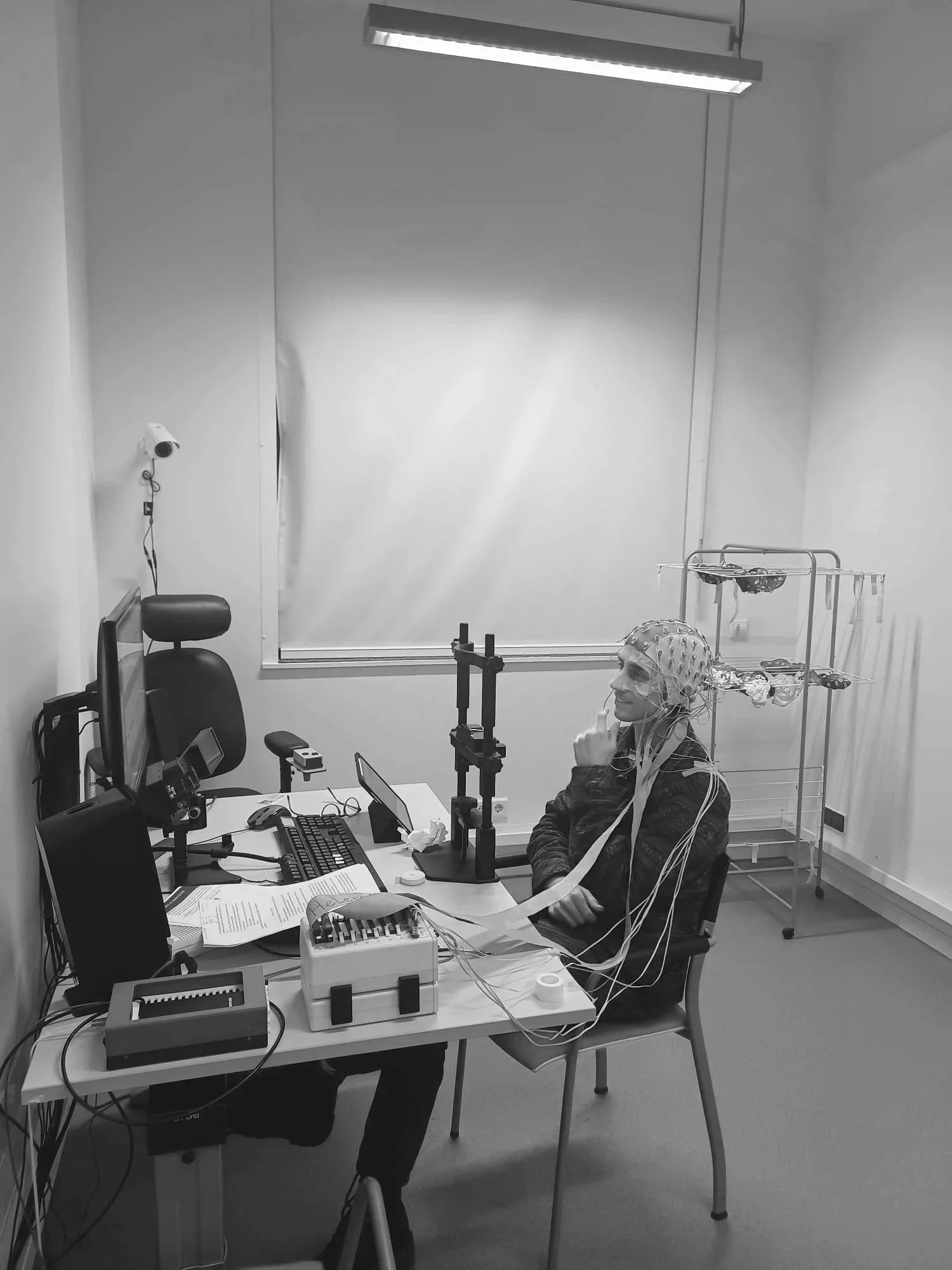 A person wearing a hospital gown and a cap with electrodes attached to their head sits in a laboratory or testing room filled with electronic equipment, wires, and monitors, with a blank white screen or window in the background.