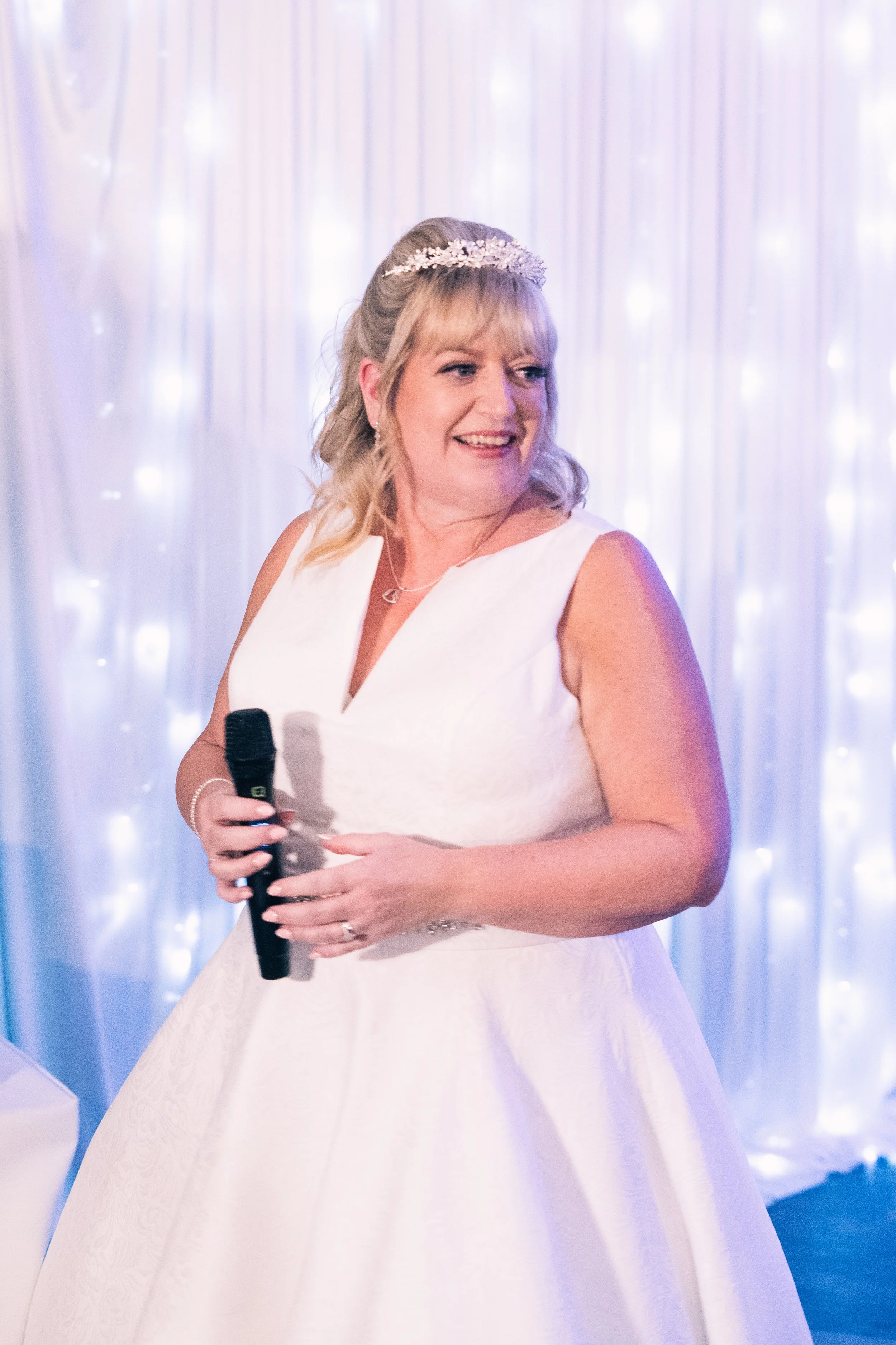 A woman in a white dress holding a microphone at a wedding reception with a lit, draped backdrop.
