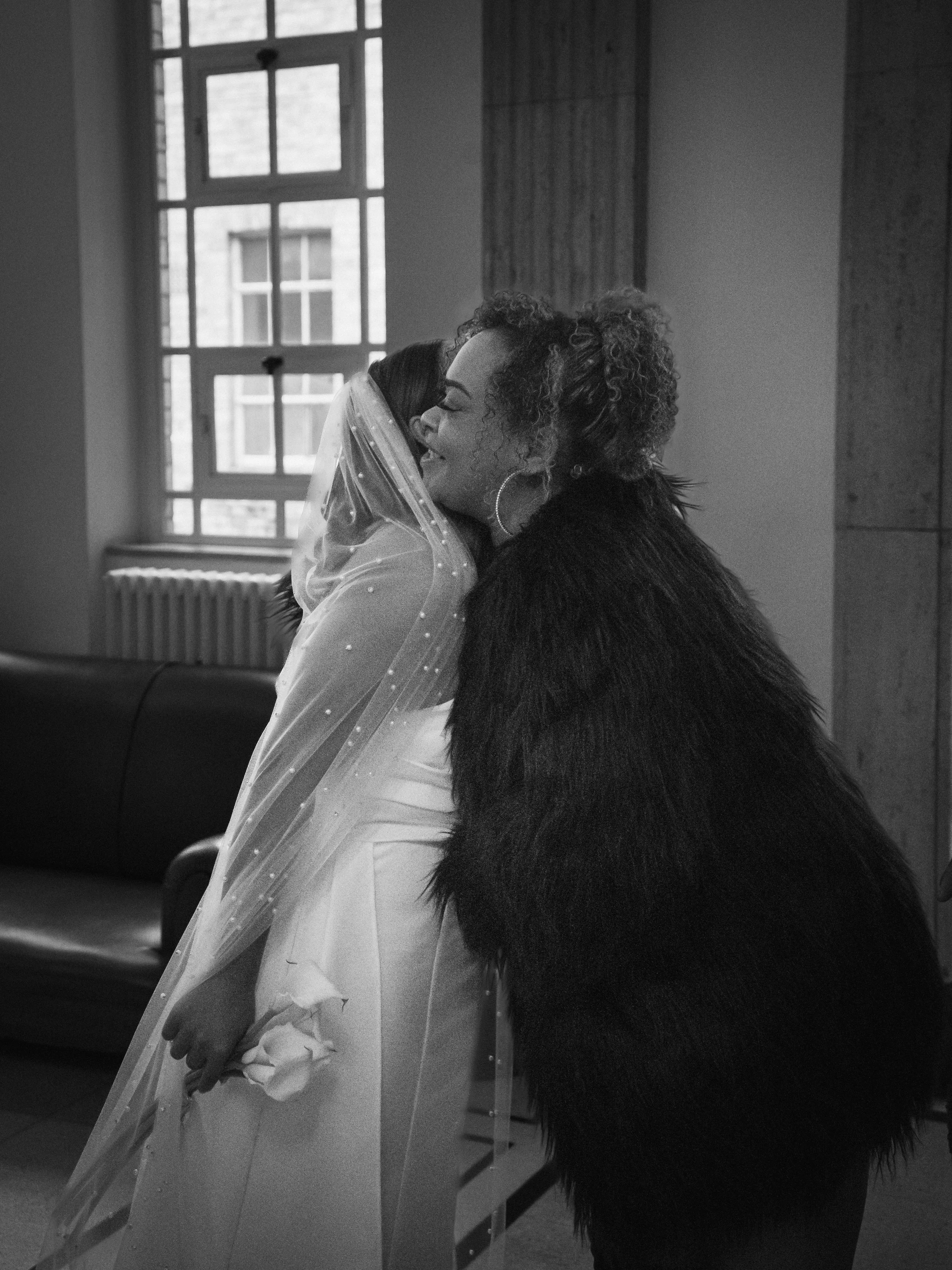 Two women embracing, one wearing a wedding dress with a veil, and the other in a black fur coat, sharing a joyful moment in a room with large windows.