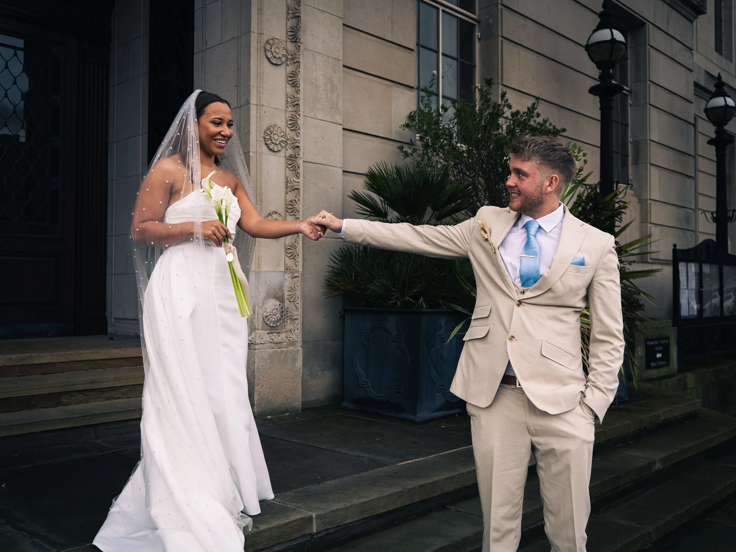 A bride and groom holding hands outside a building, smiling at each other. The bride is in a white wedding gown with a veil and holding a bouquet. The groom is in a tan suit with a blue tie and pocket square.