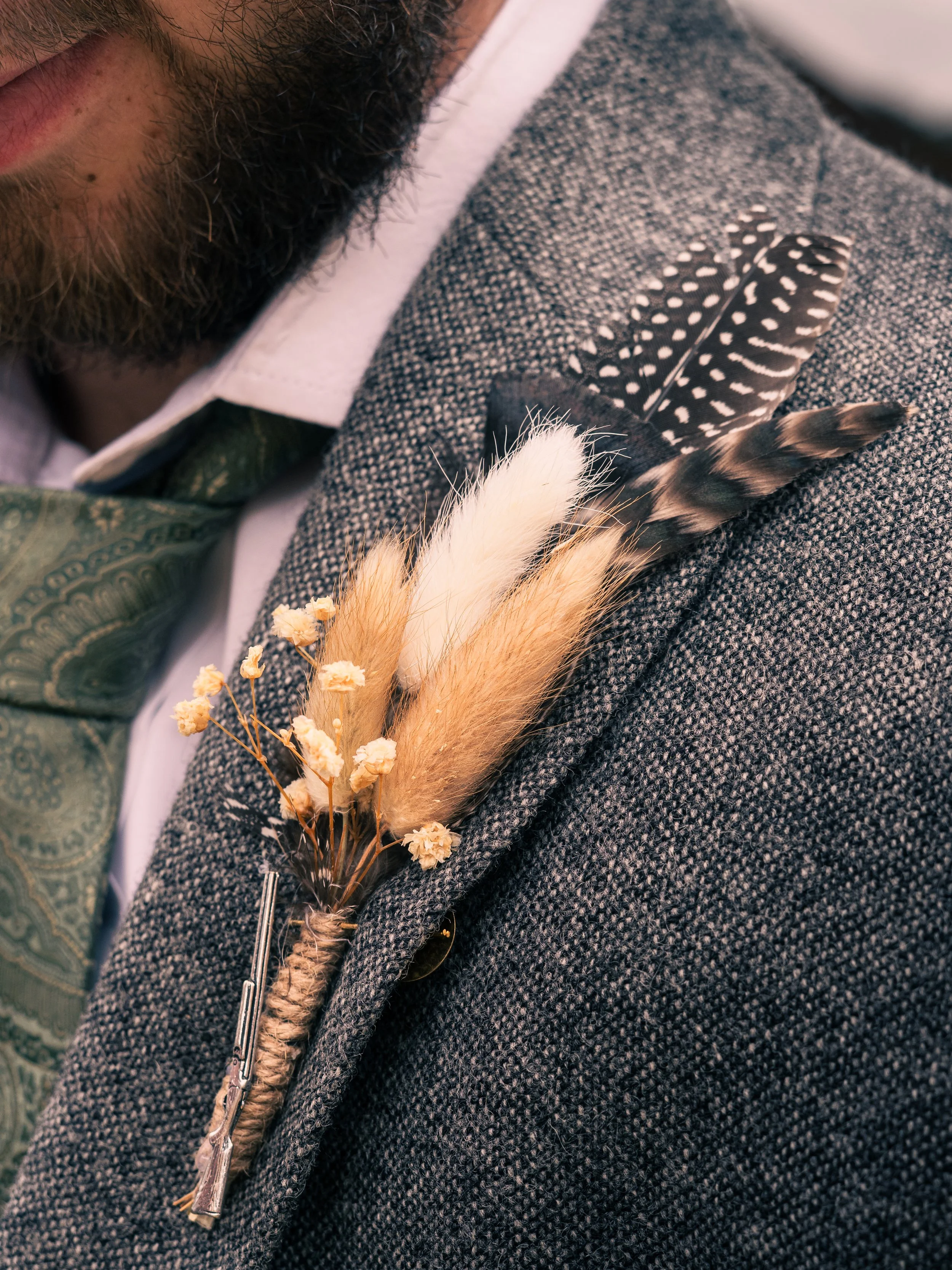 Close-up of a man's suit jacket with a boutonniere made of feathers and dried flowers attached to the lapel.
