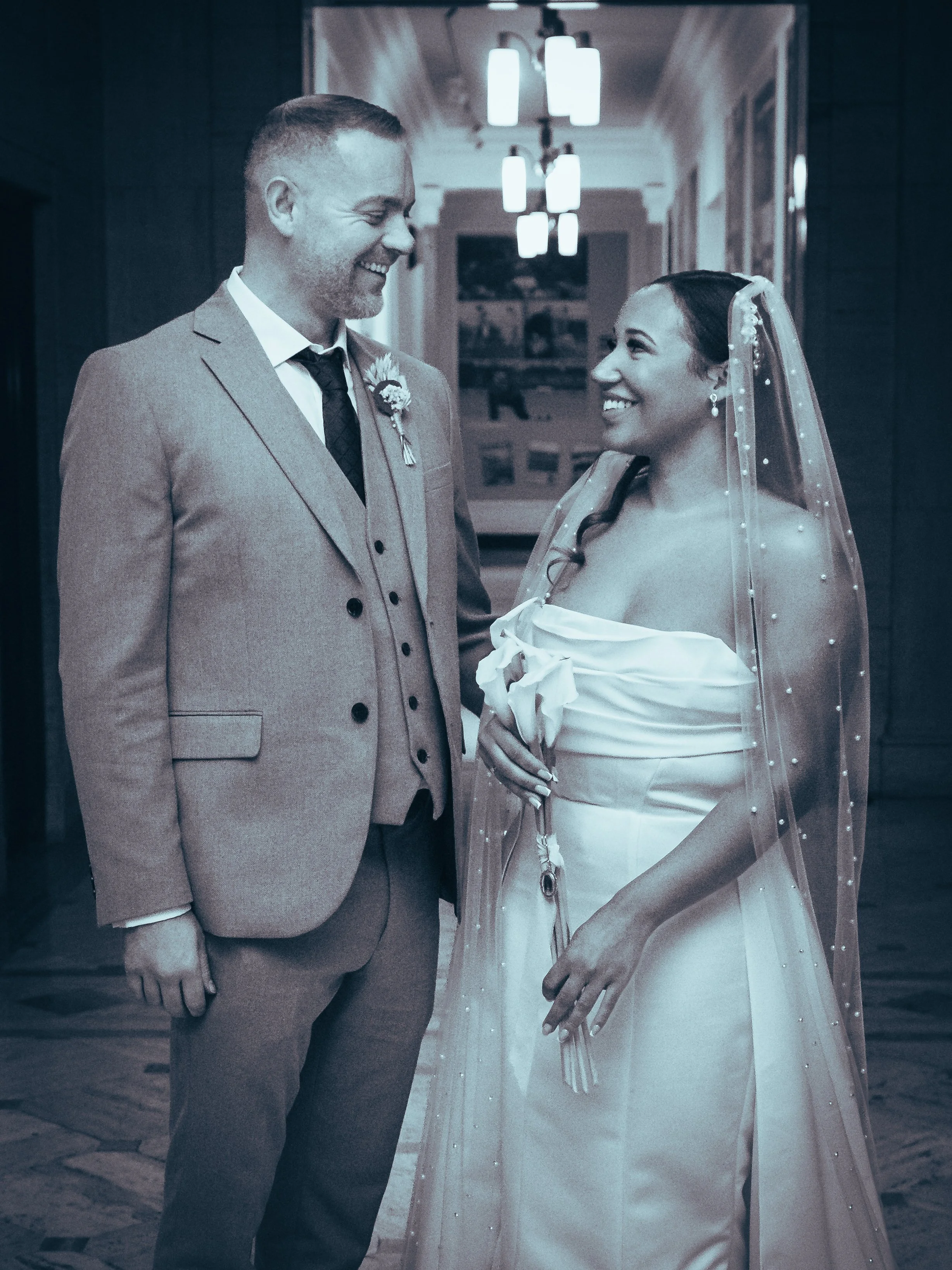 A couple on their wedding day, the groom in a light-colored suit and the bride in a strapless wedding gown with a veil, standing face to face indoors, smiling at each other.