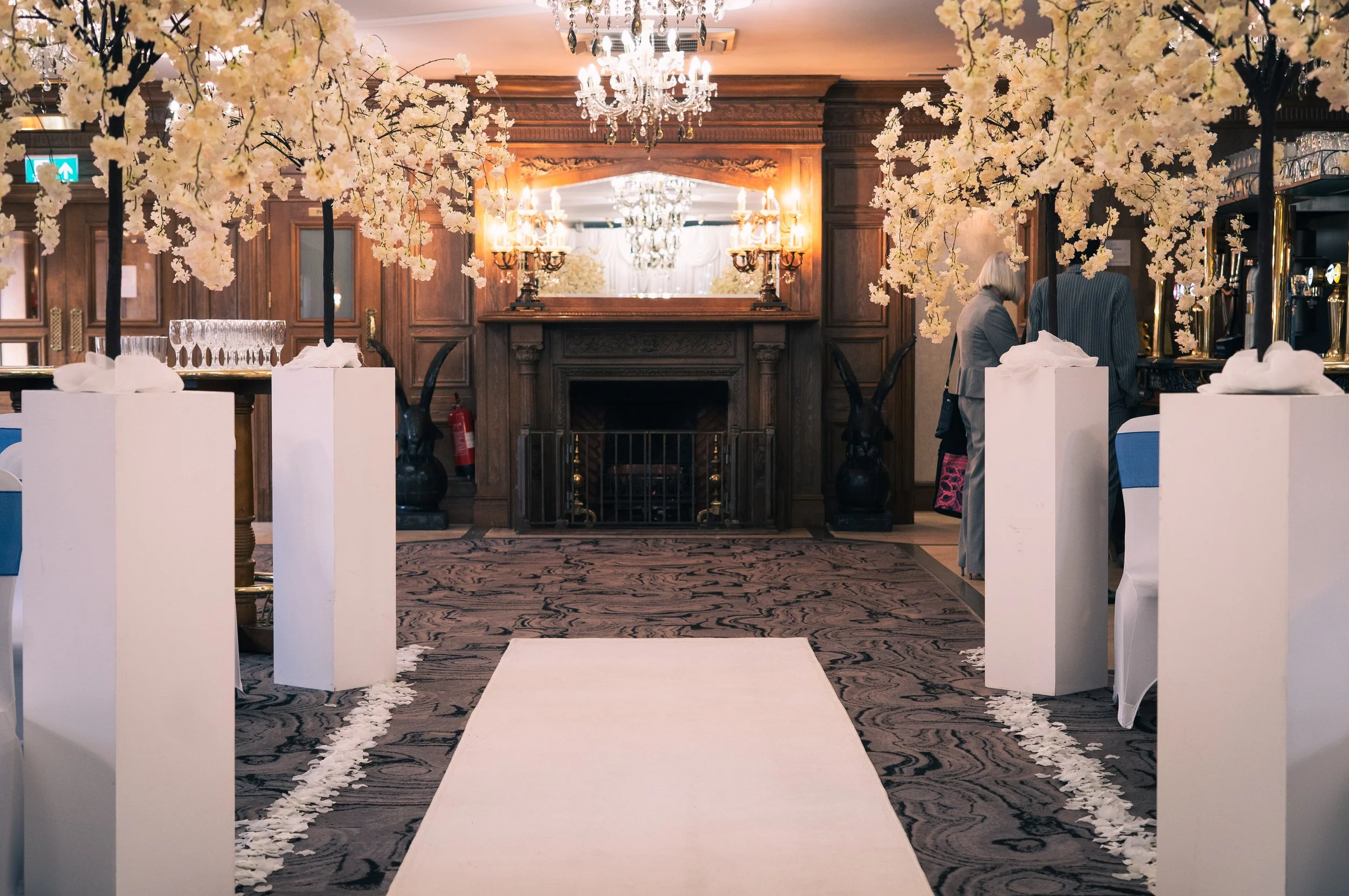 Elegant wedding ceremony setup with a white aisle runner, surrounded by tall white floral arrangements on stands, in a room with wooden paneling, chandeliers, and a fireplace in the background.