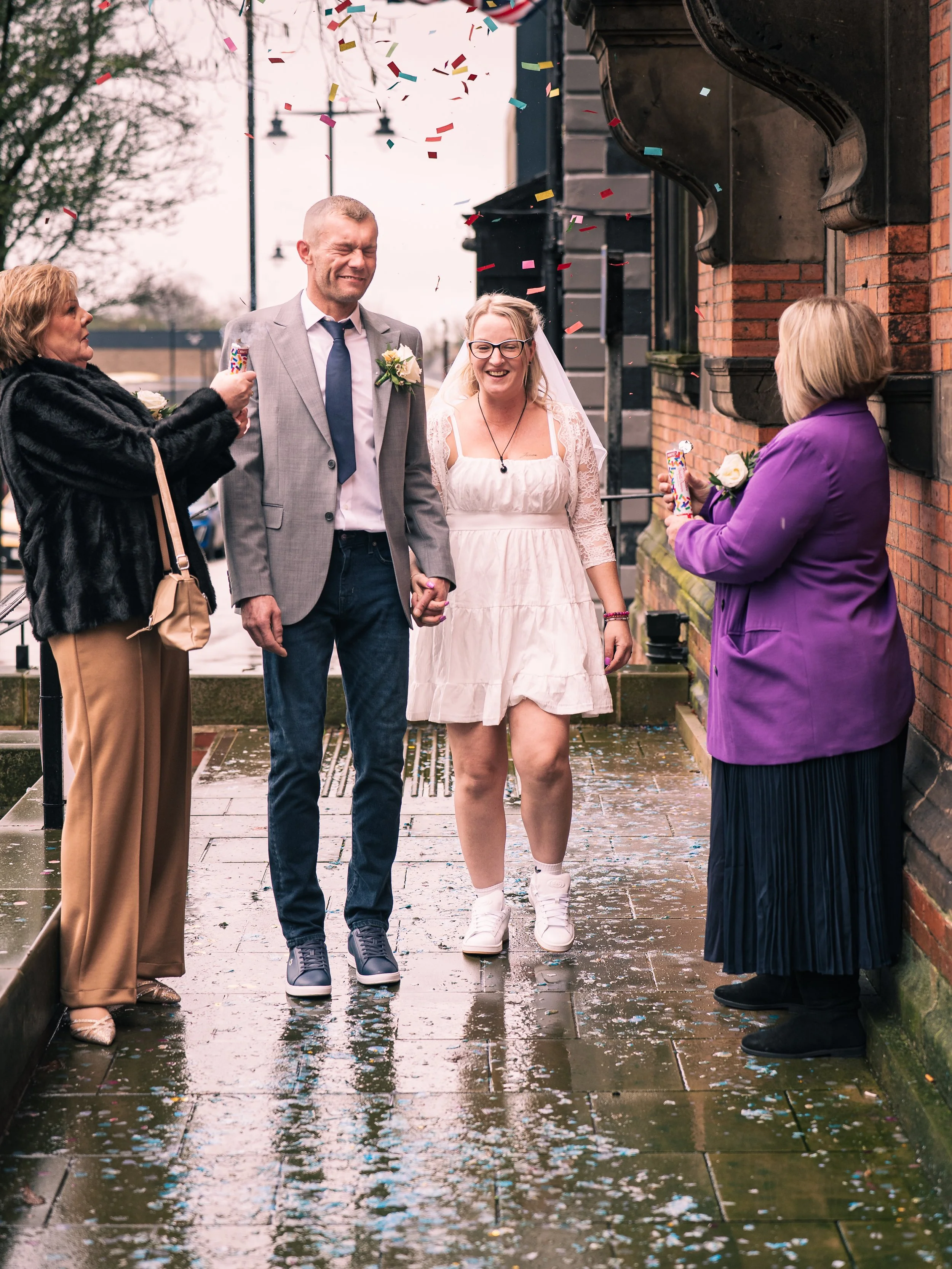 A bride and groom holding hands walking outdoors on a rainy day, celebrating with family friends, with confetti falling in the air.