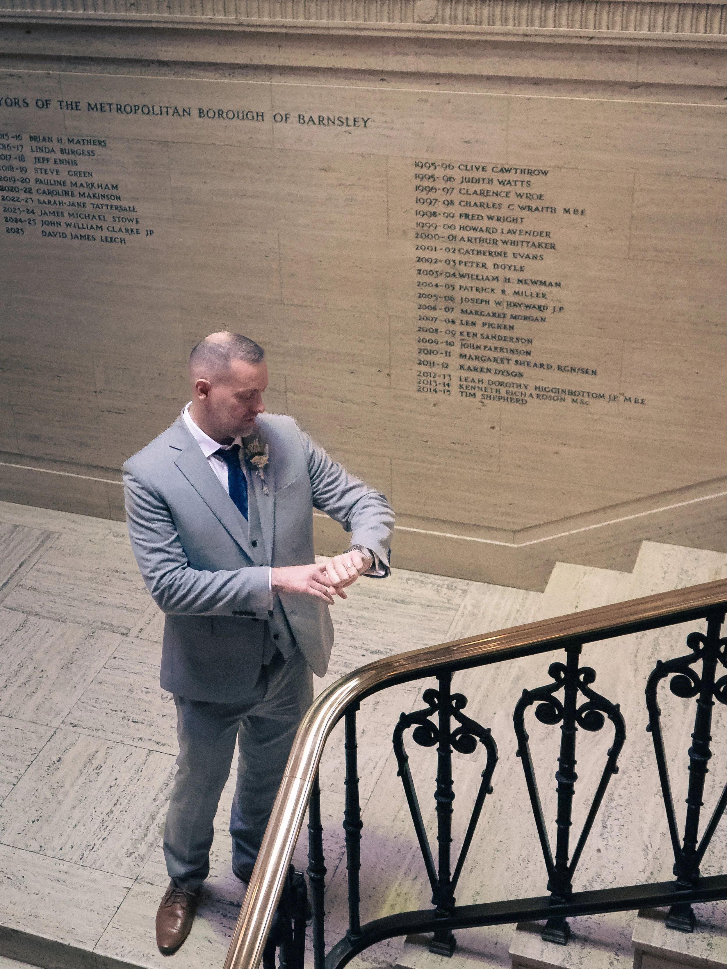 A man in a light gray suit looking at his watch on a staircase landing in an interior space with a memorial wall in the background listing names and years, possibly a memorial or honor wall.