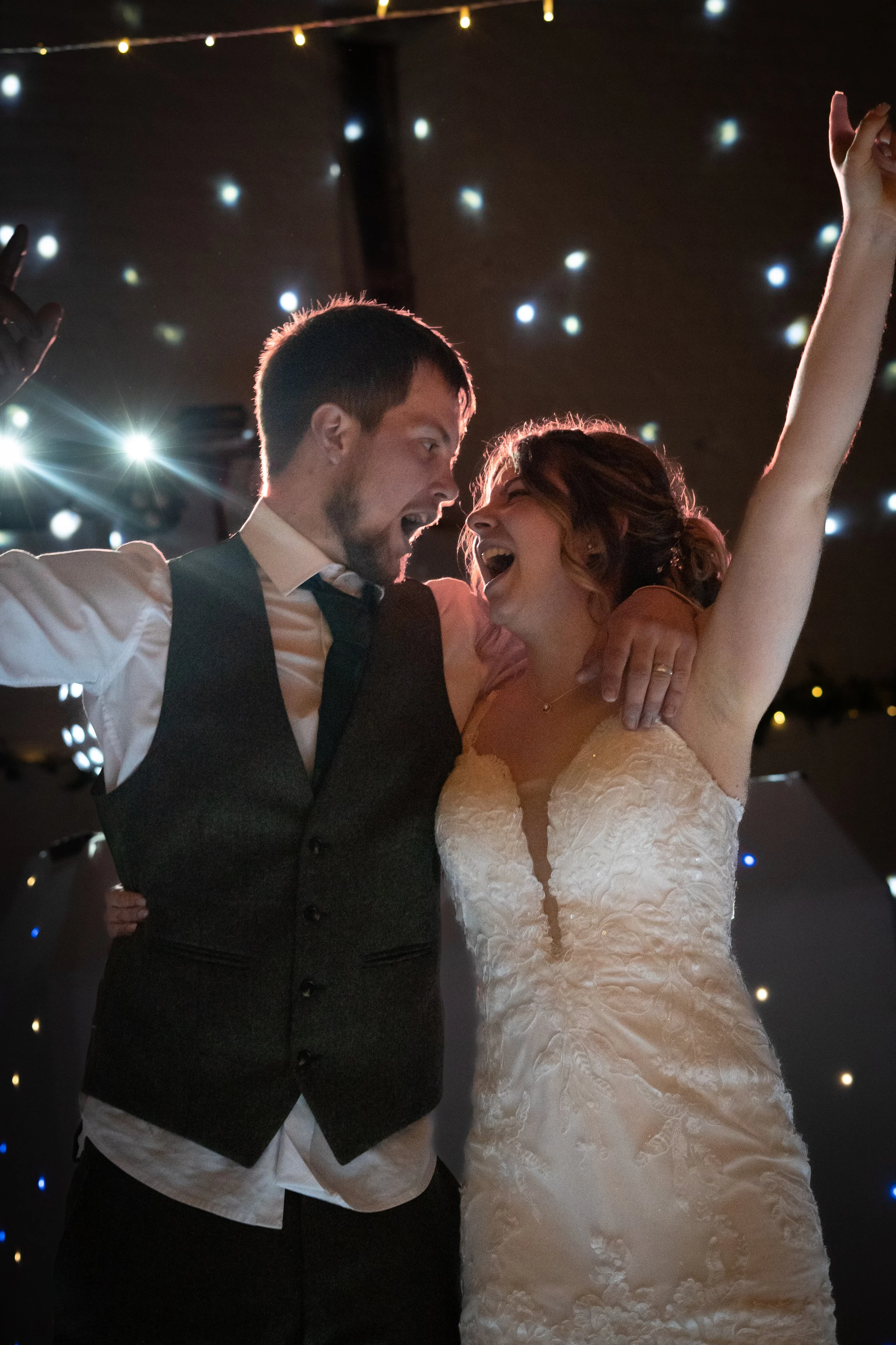 A newlywed couple dancing and celebrating, with the groom singing and the bride smiling with her arm raised, in a festive setting with string lights and star-like decorations.
