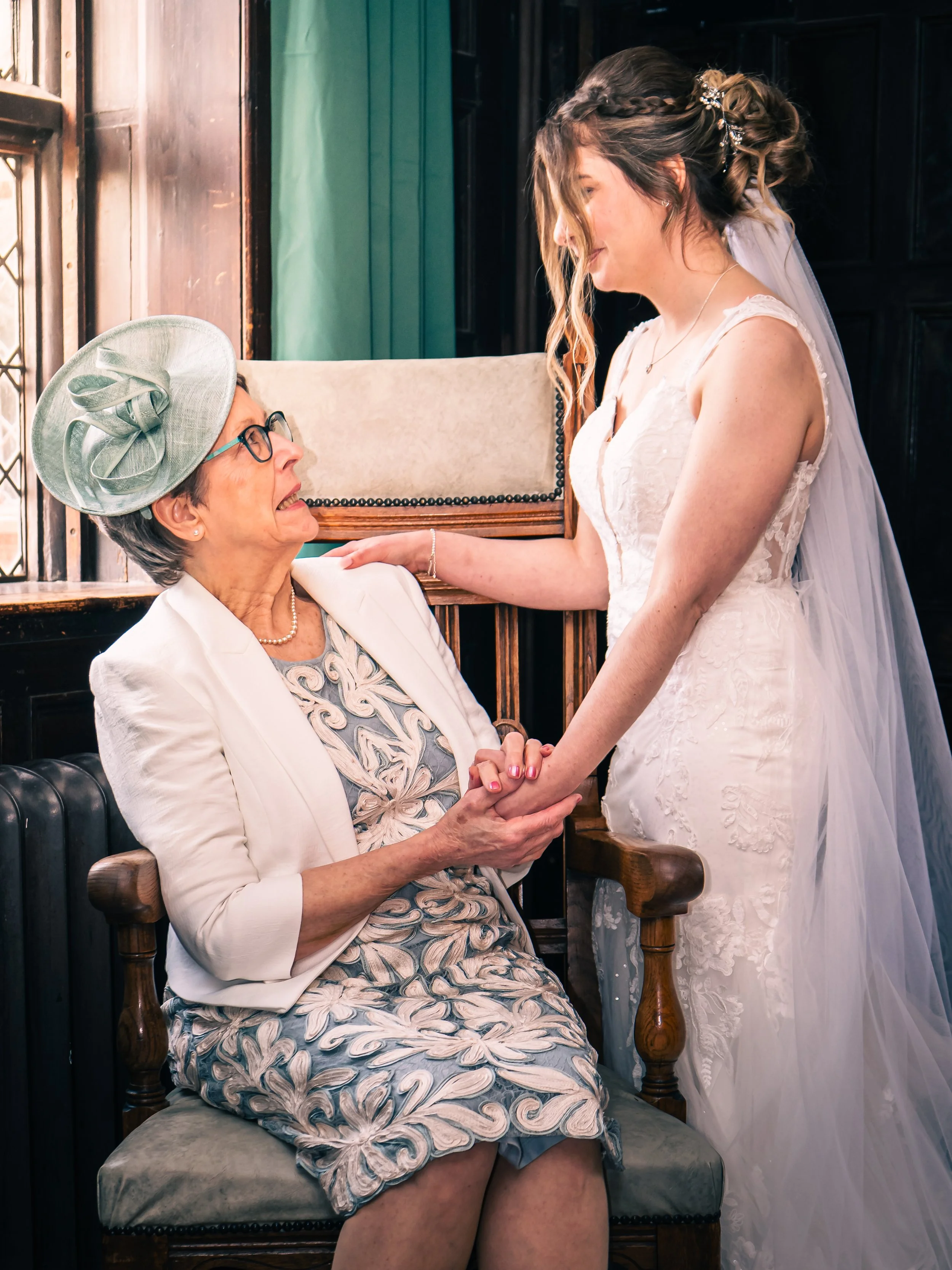 A bride with long, wavy hair in a braided half-up style, wearing a white lace wedding gown with a veil, holding hands with an elderly woman in a floral dress, white blazer, light green hat with a bow, and glasses, inside a warmly lit room with dark w