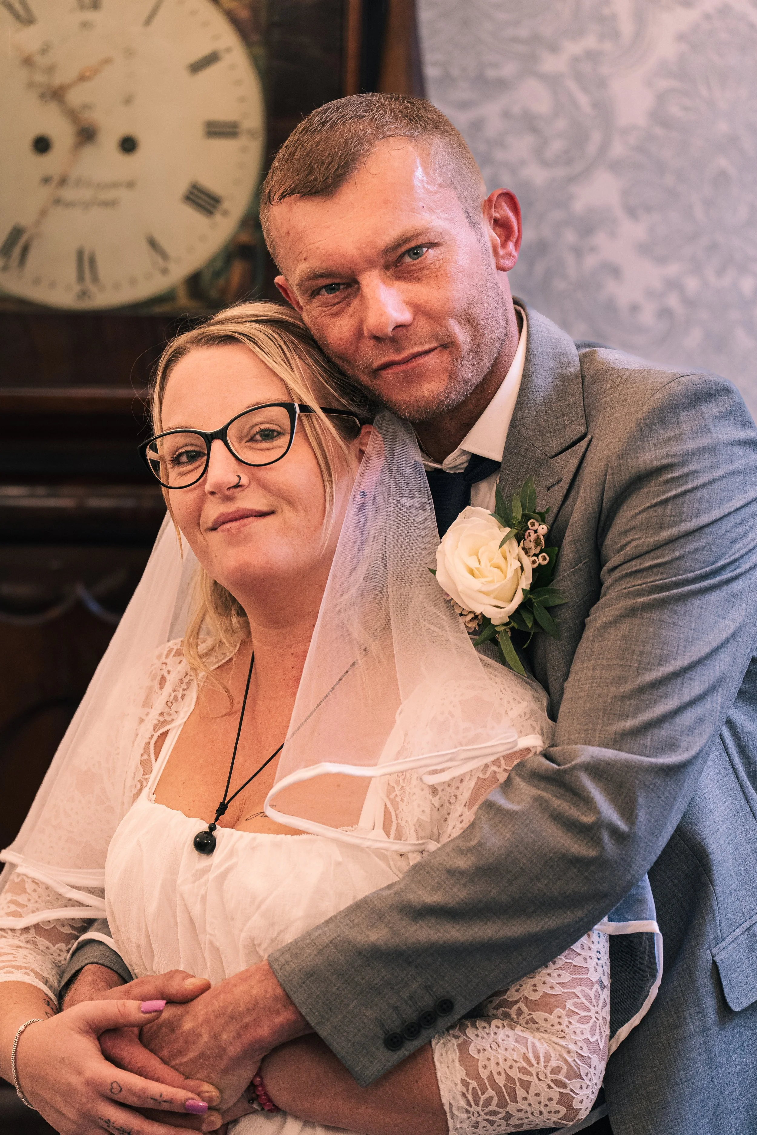 A bride and groom hugging at their wedding, with the bride wearing glasses, a white lace dress, and a veil, and the groom in a gray suit with a white rose boutonniere.