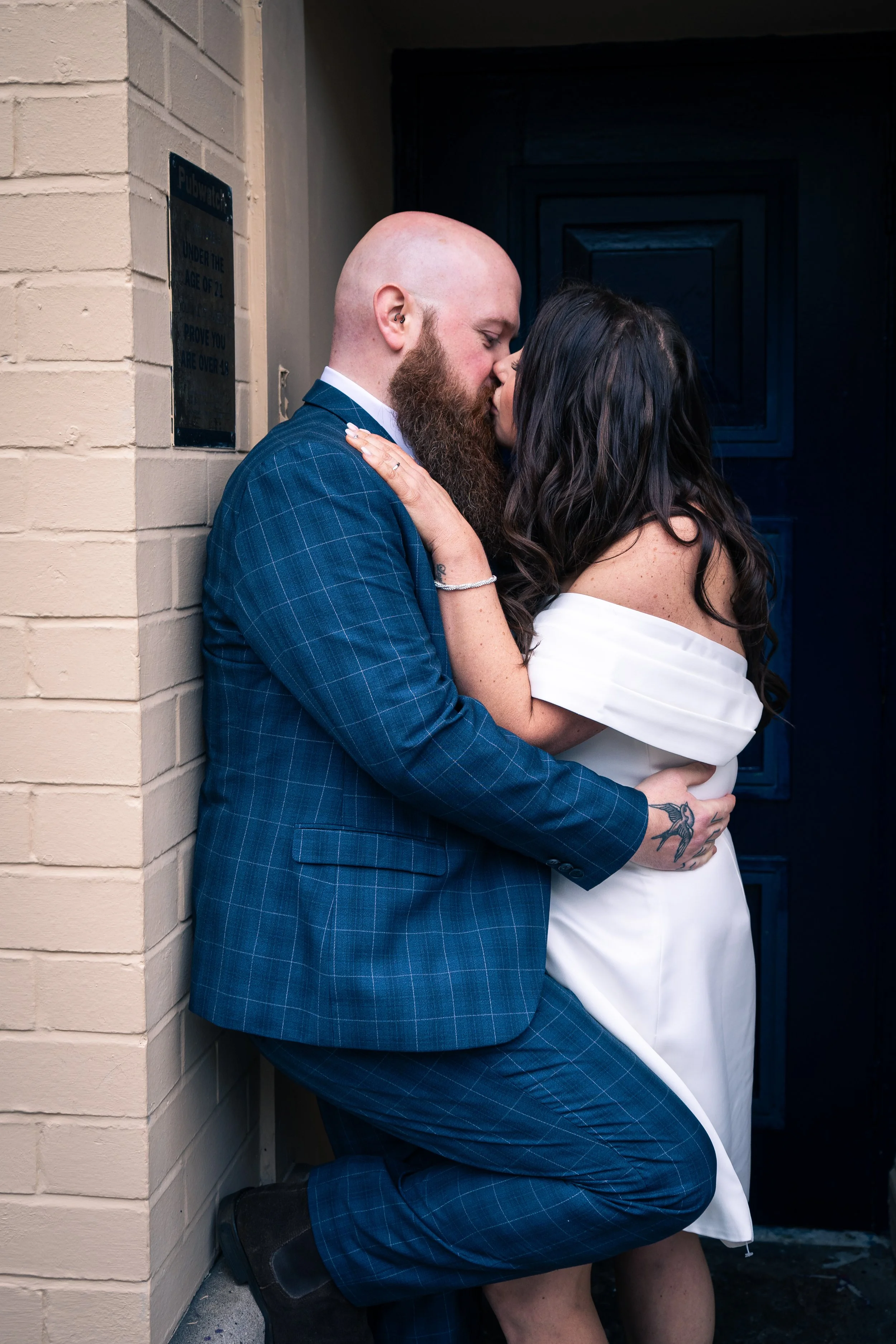 A man in a blue checkered suit and a woman in an off-shoulder white dress sharing a kiss while leaning against a beige brick wall and a dark door.