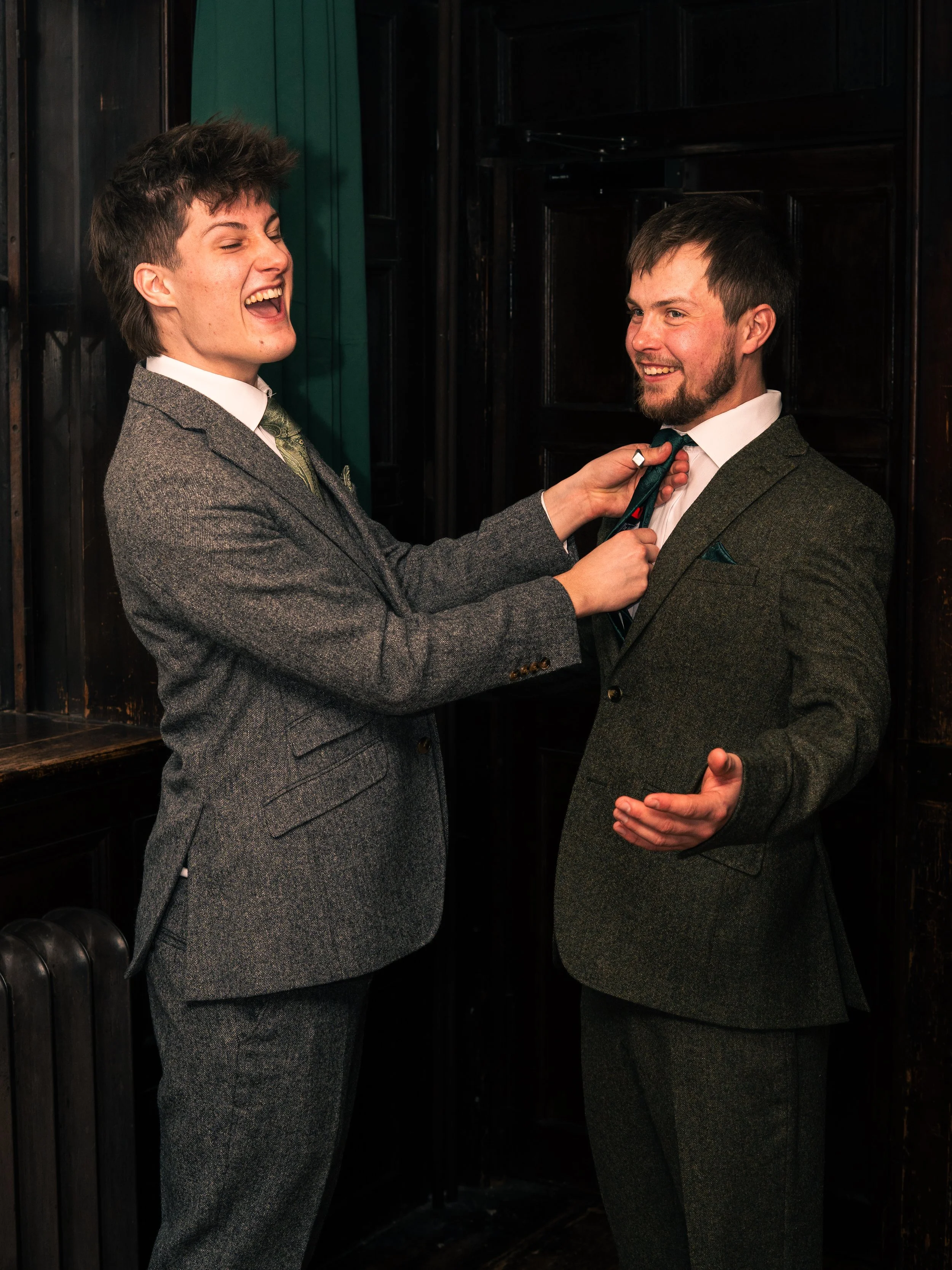 Two men in suits smiling and laughing, one adjusting the other's tie in a dimly lit room with dark wood paneling.