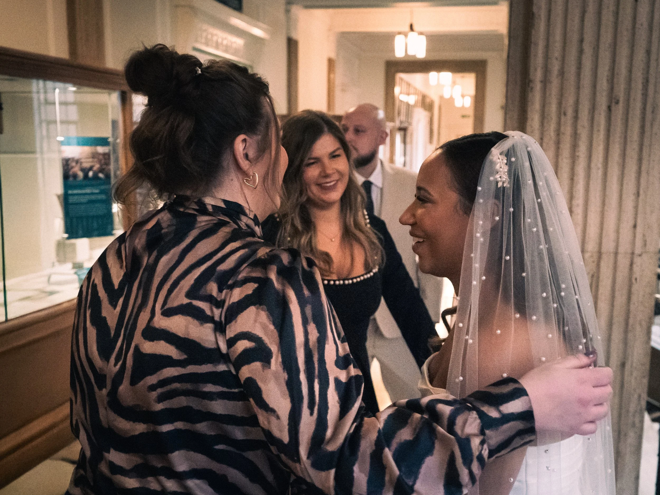 Two women, one wearing a patterned jacket and the other a wedding dress with a veil, are smiling and holding hands during a wedding celebration. There are three other people, two women and one man, in the background smiling and engaging in conversati