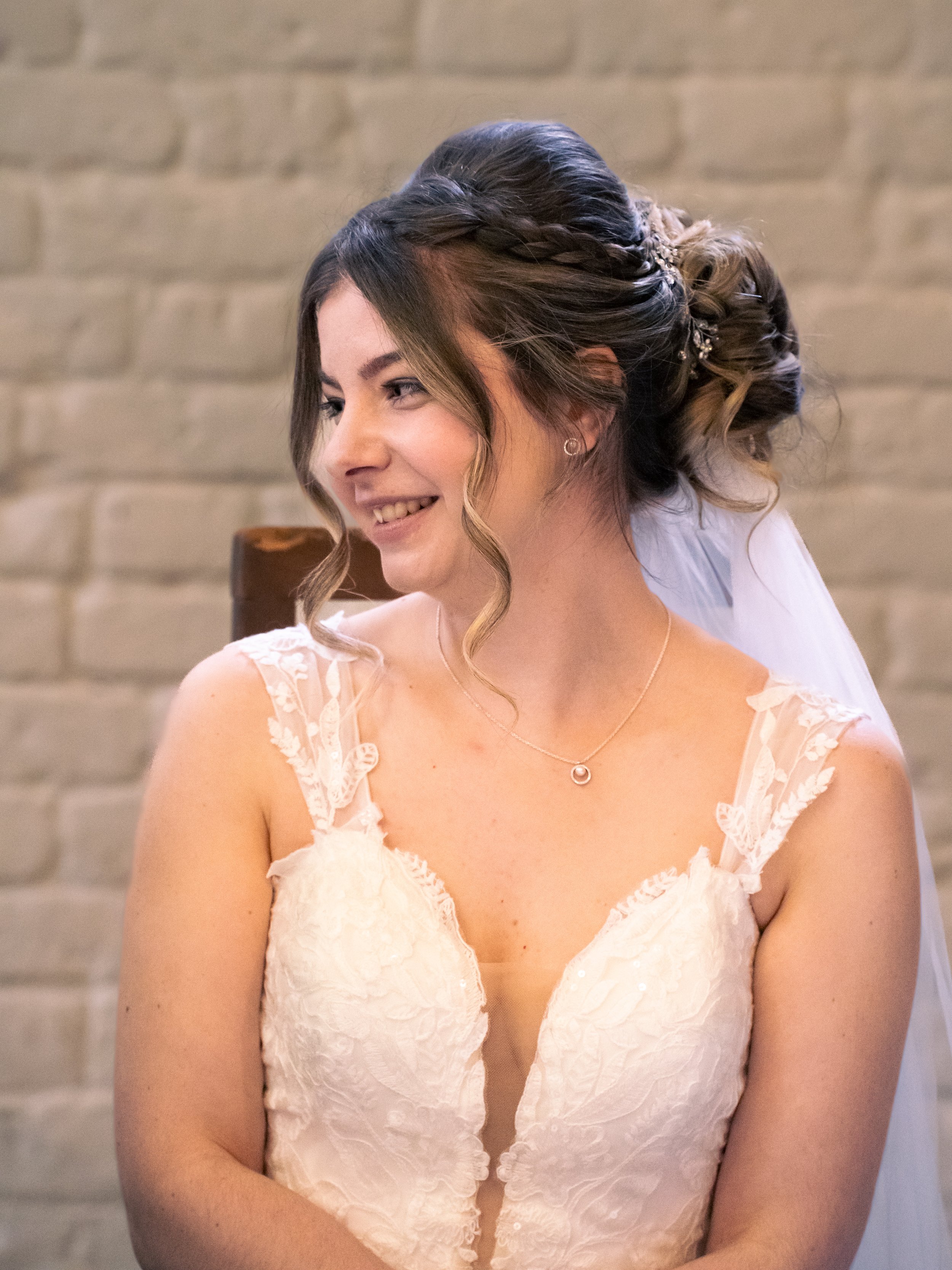 A smiling bride with dark brown hair styled in an elegant updo with loose curls and a decorative hairpiece, wearing a lace wedding dress with a deep V neckline and delicate shoulder straps, and a simple necklace, standing in front of a light brick wa