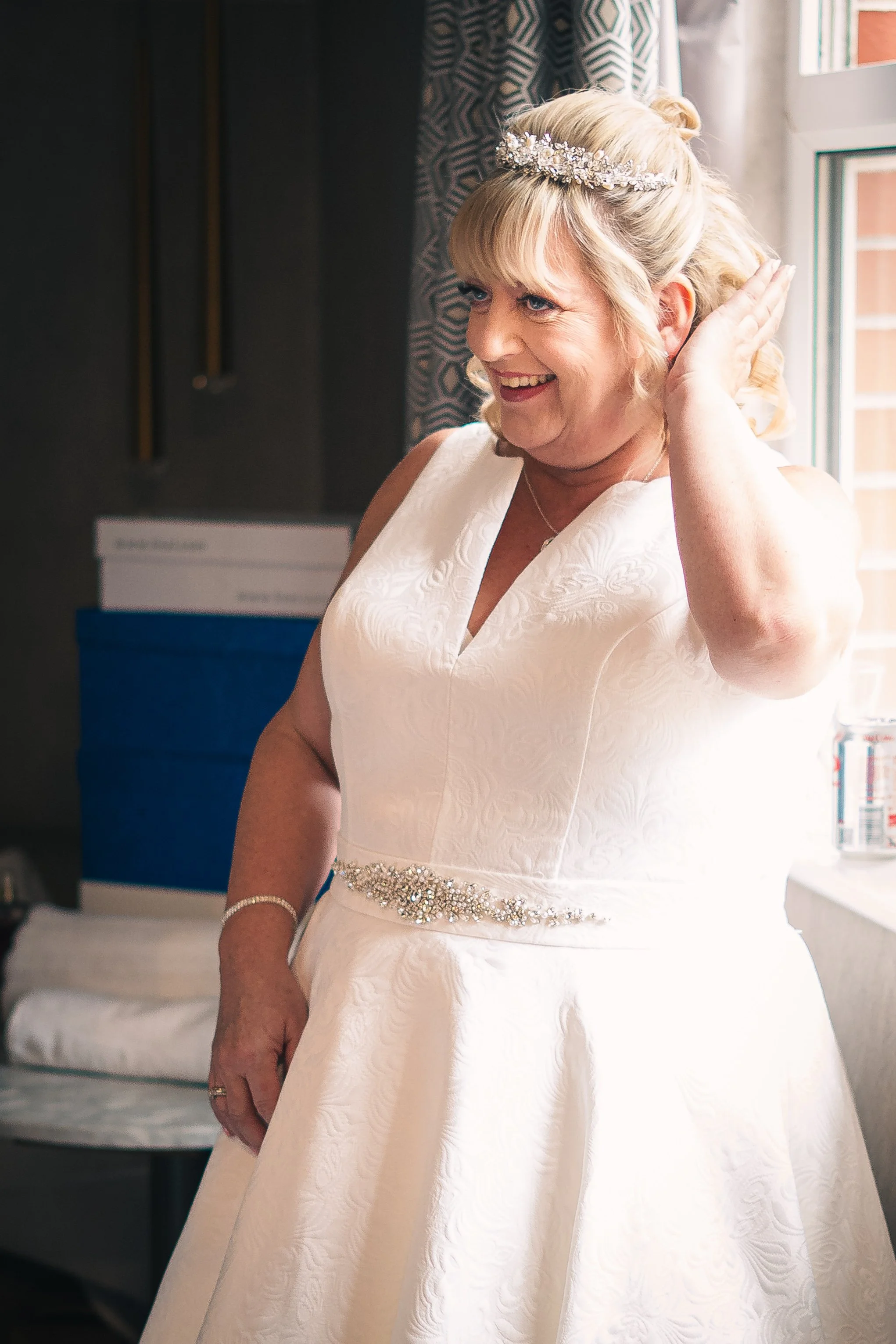 A happy woman in a white wedding dress with a bejeweled belt, wearing a tiara, smiling as she touches her hair.