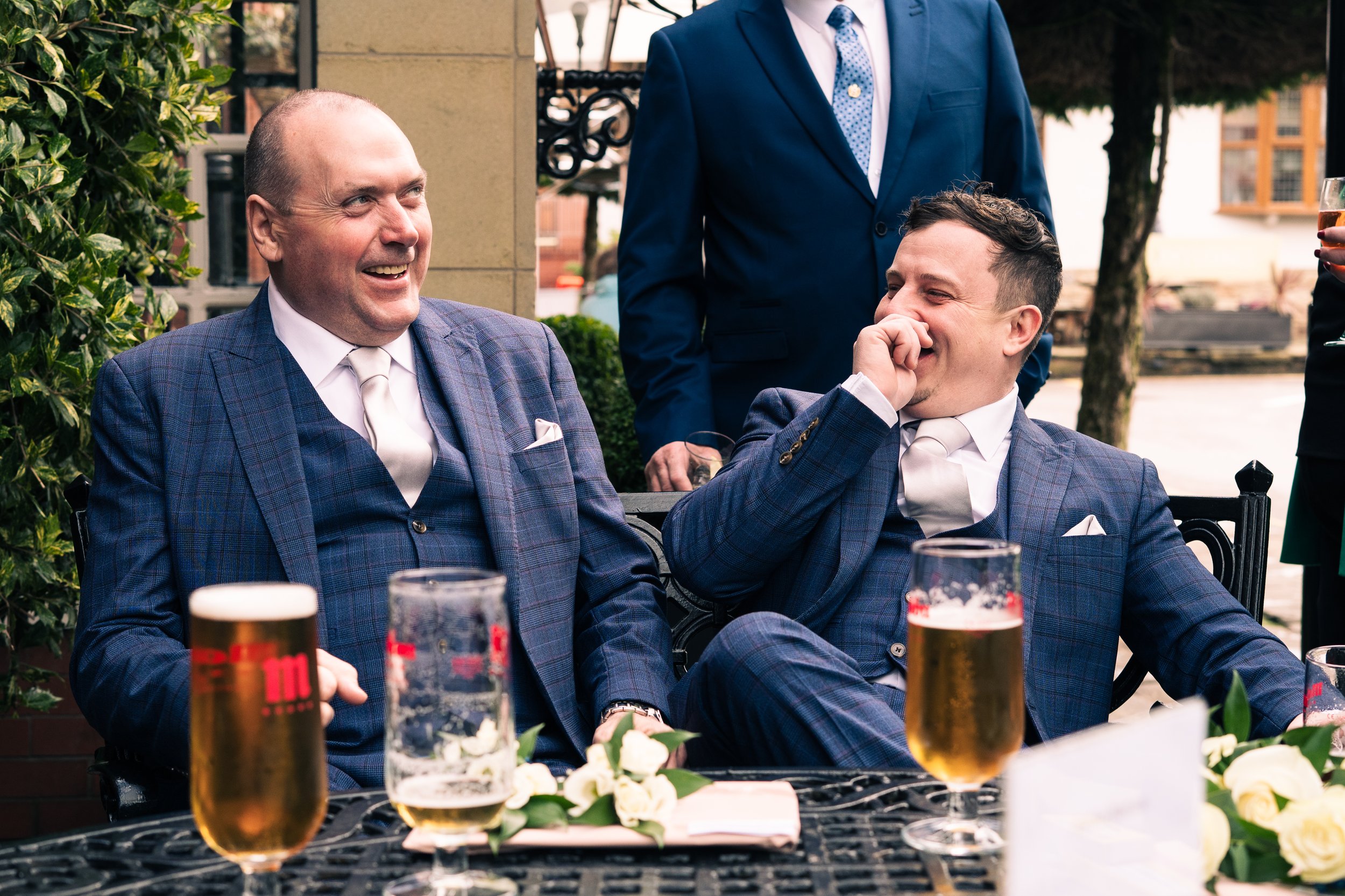Two men in blue suits sitting at a table and laughing, with drinks in front of them, during an outdoor celebration or wedding