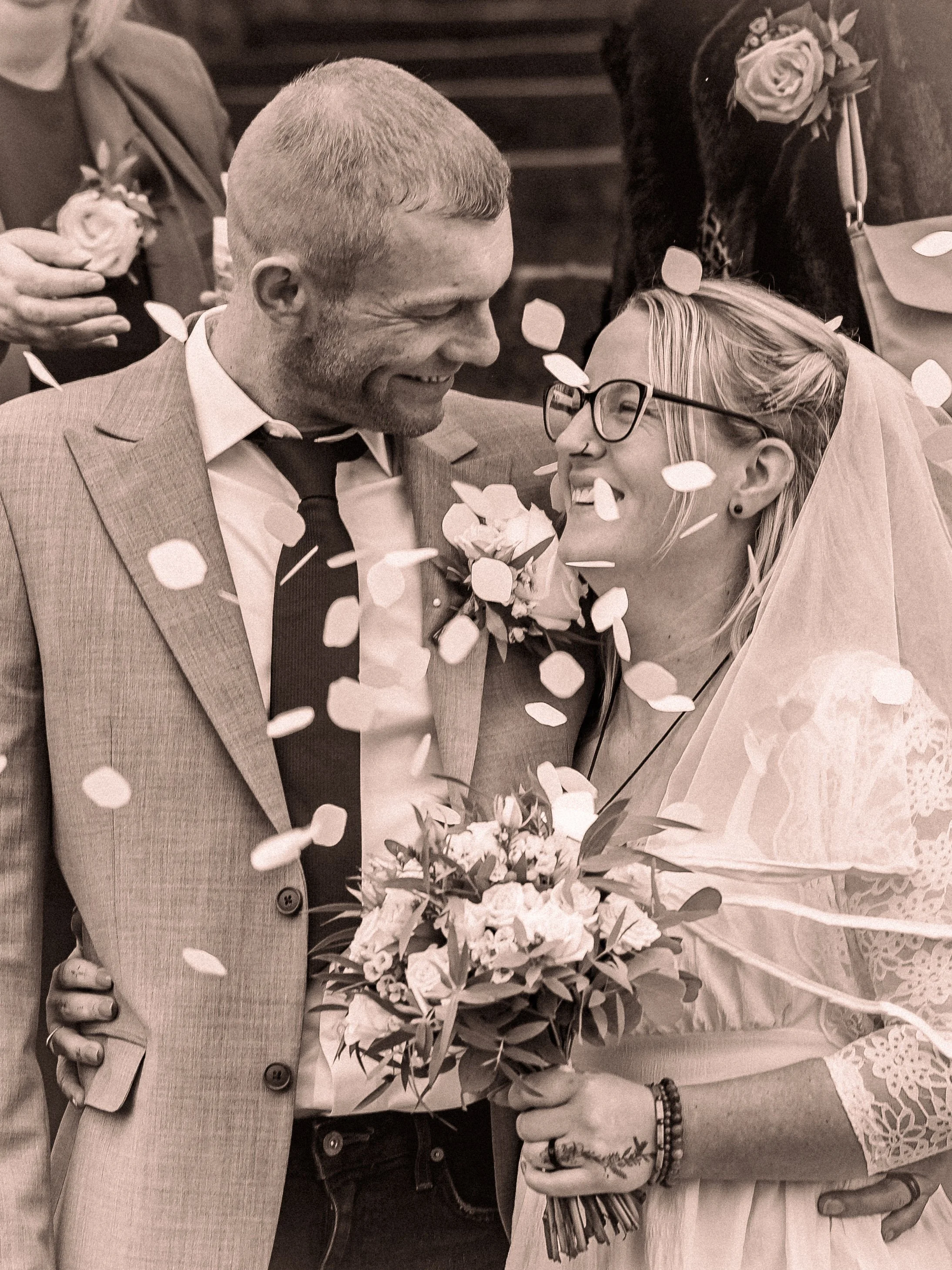 A couple at a wedding reception, smiling and looking into each other's eyes, with a man wearing a suit and a woman wearing glasses and a veil, holding a bouquet. Confetti is falling around them.