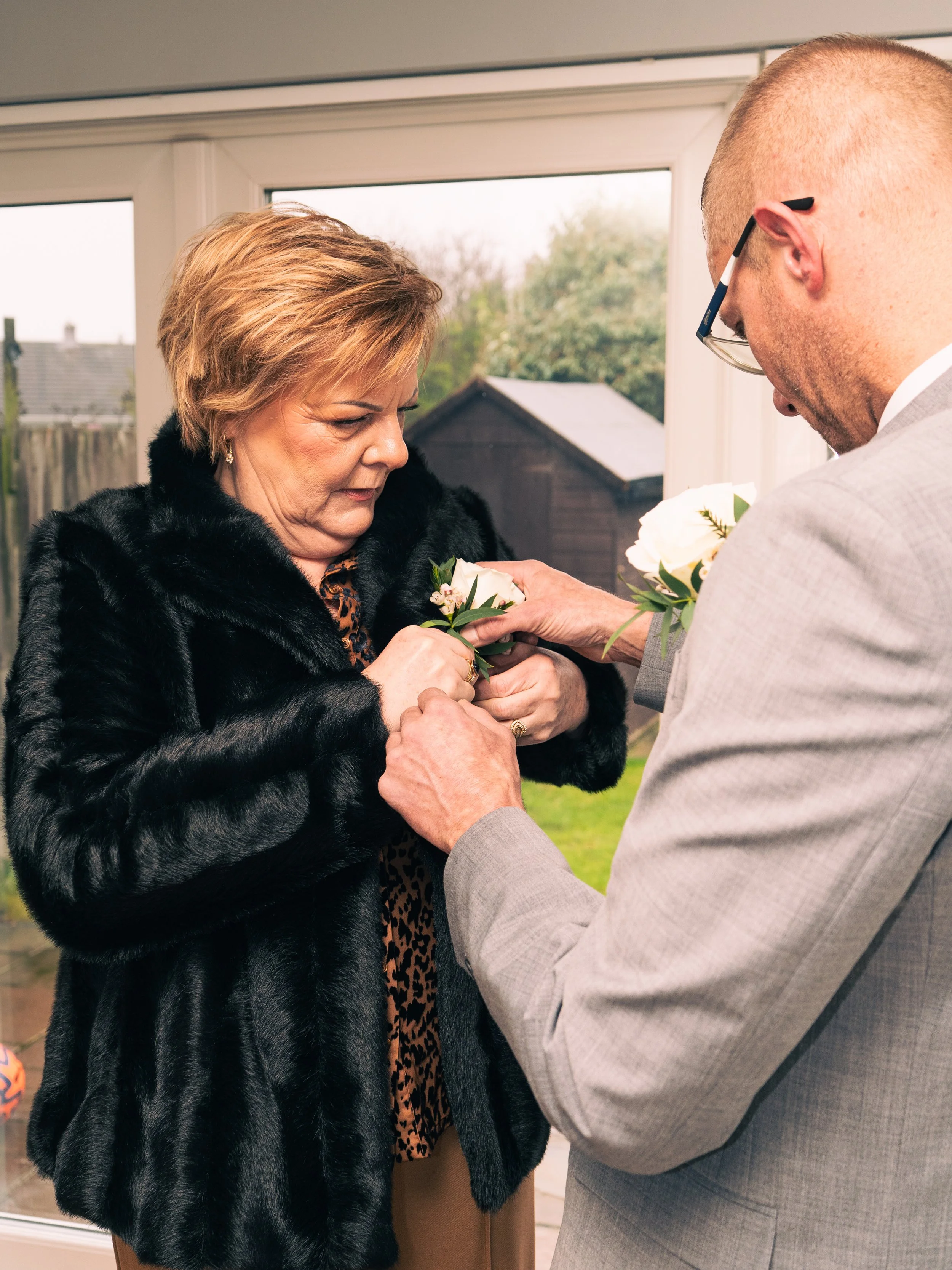 A man in a gray suit pins a corsage onto a woman's clothing inside a house, with a window showing a backyard in the background.