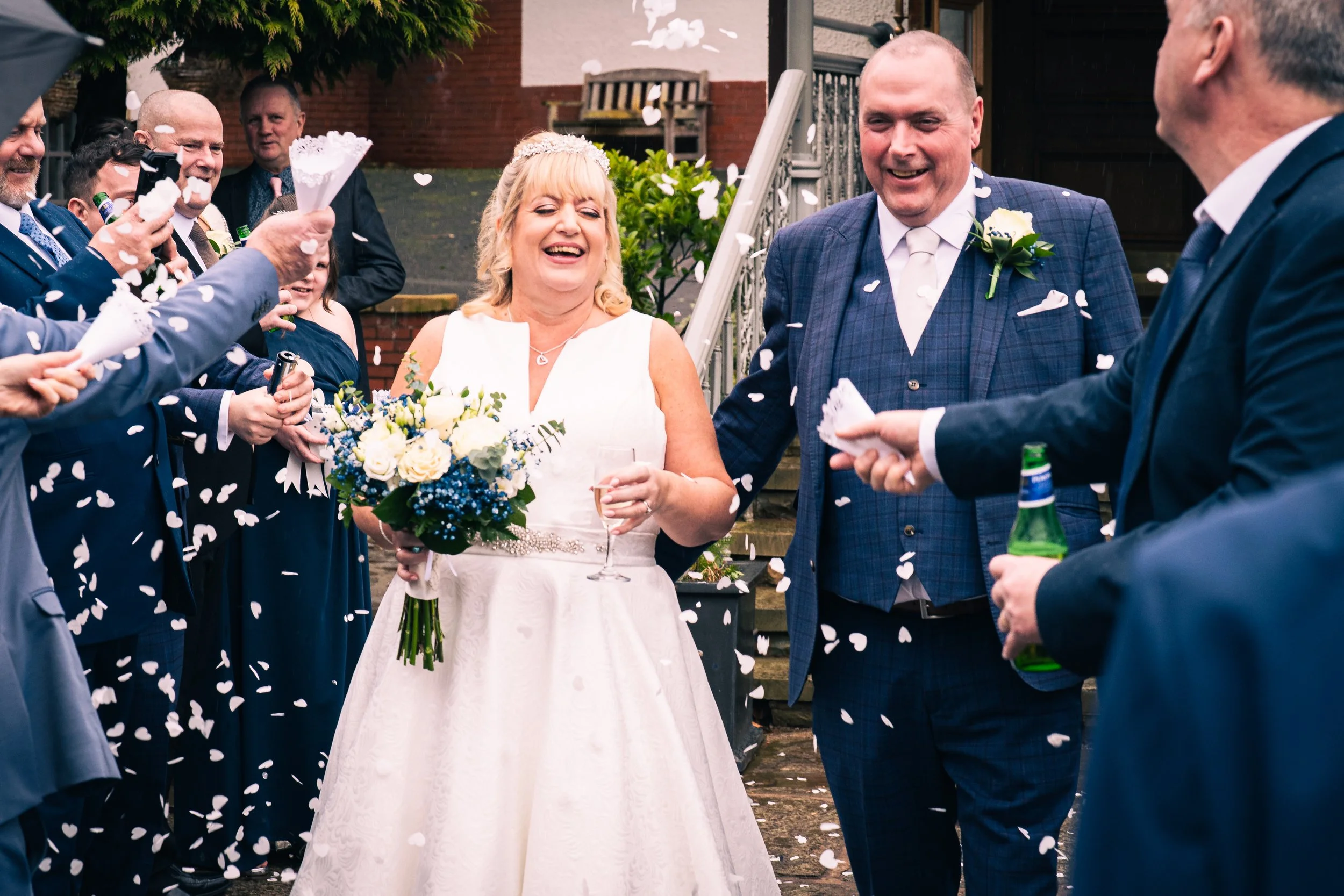 A wedding celebration outdoors with a happy bride holding a bouquet and a glass of champagne, standing next to a smiling groom in a suit, surrounded by guests throwing confetti.