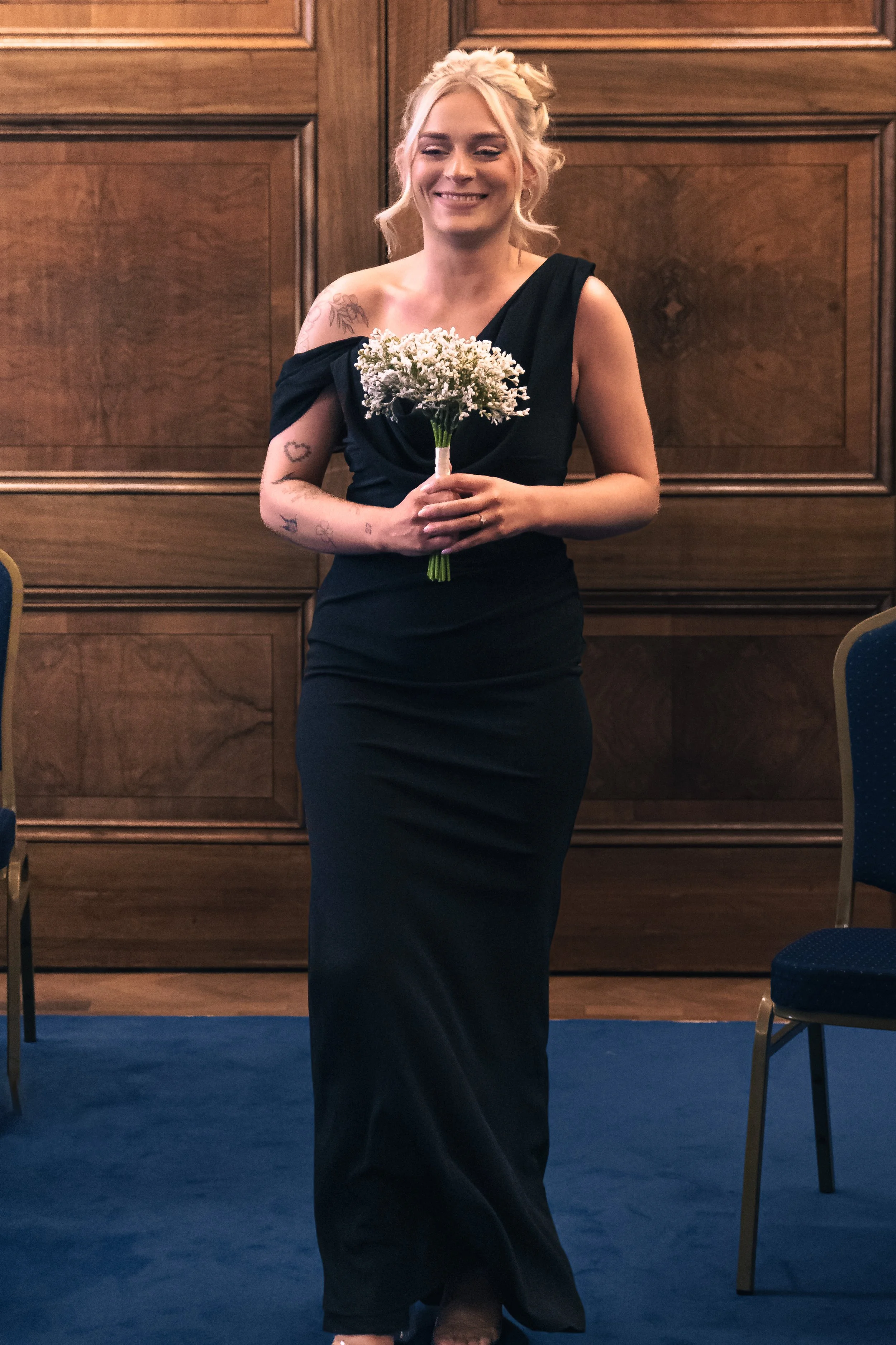 A woman in a long black dress holding a bouquet of white flowers, standing in a room with wooden wall paneling and blue carpeted floor.