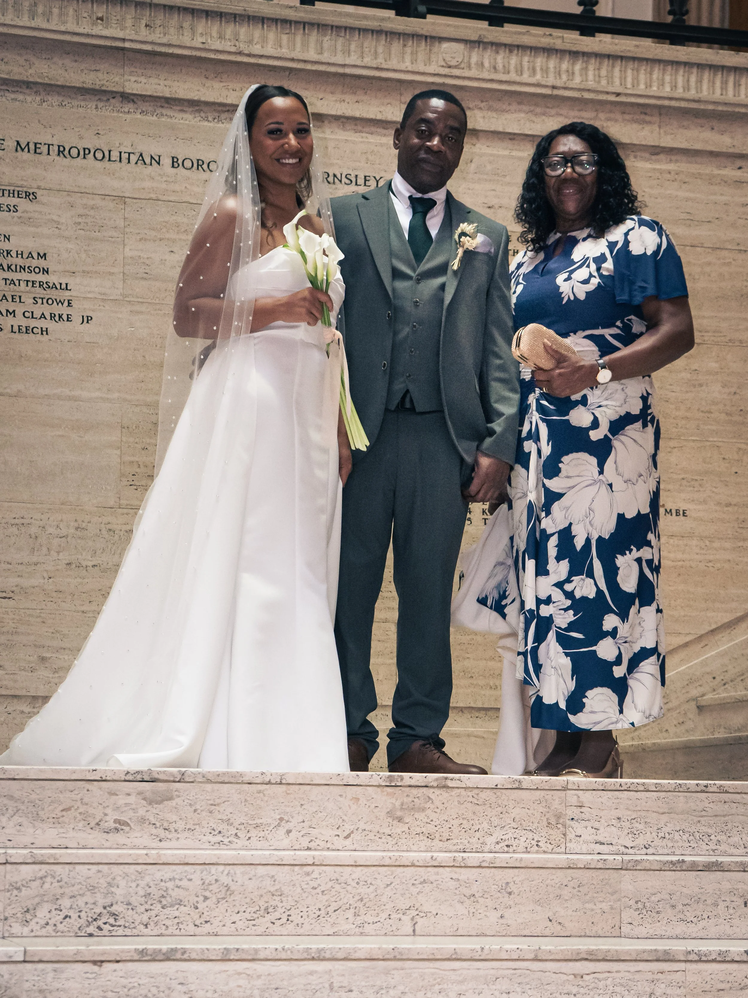 A bride in a white wedding dress holding a bouquet stands beside a groom in a gray suit, with her mother in a blue floral dress and glasses at a wedding ceremony.