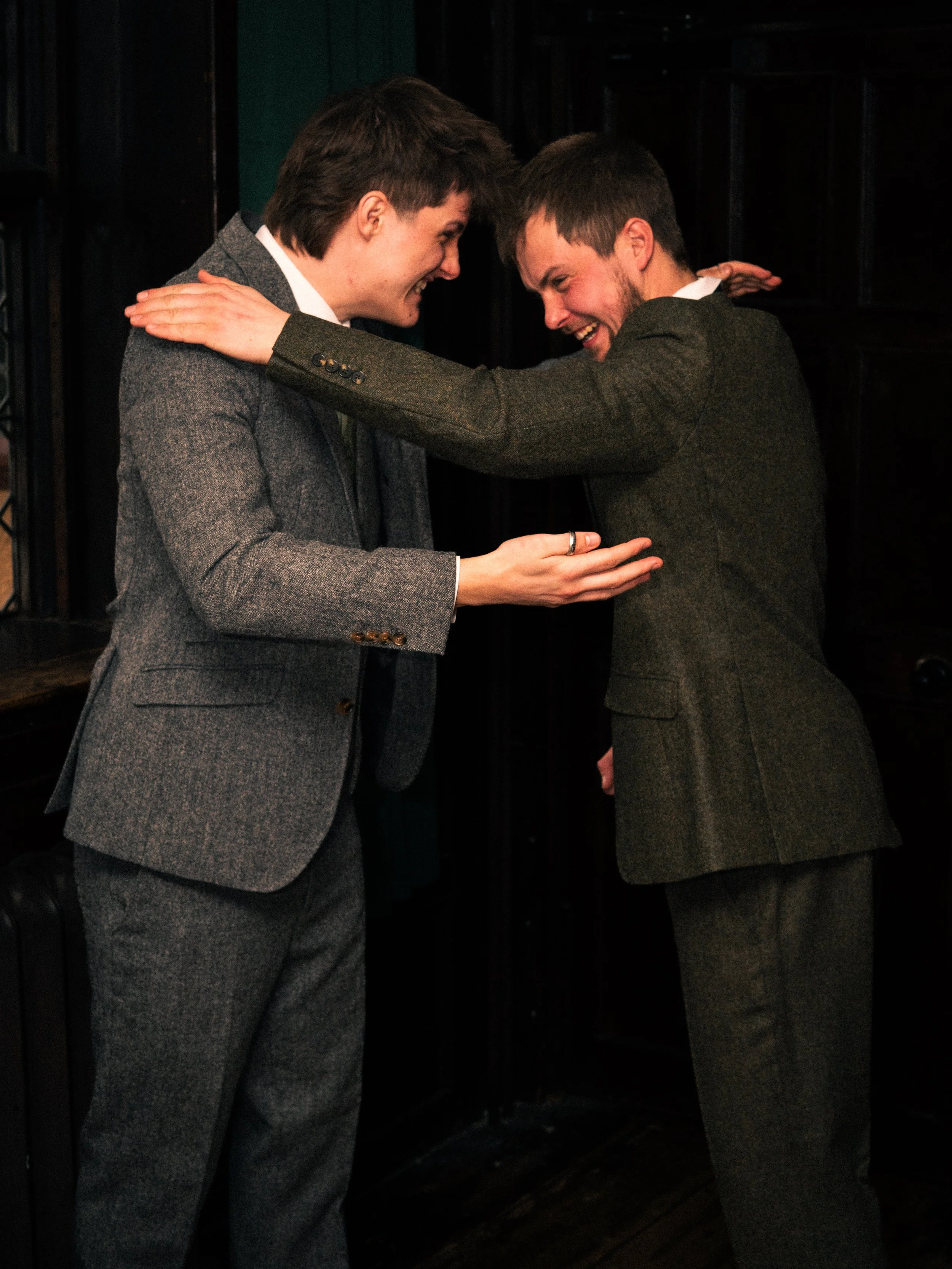 Two men in suits sharing a joyful hug and smiling at each other in a dark room.