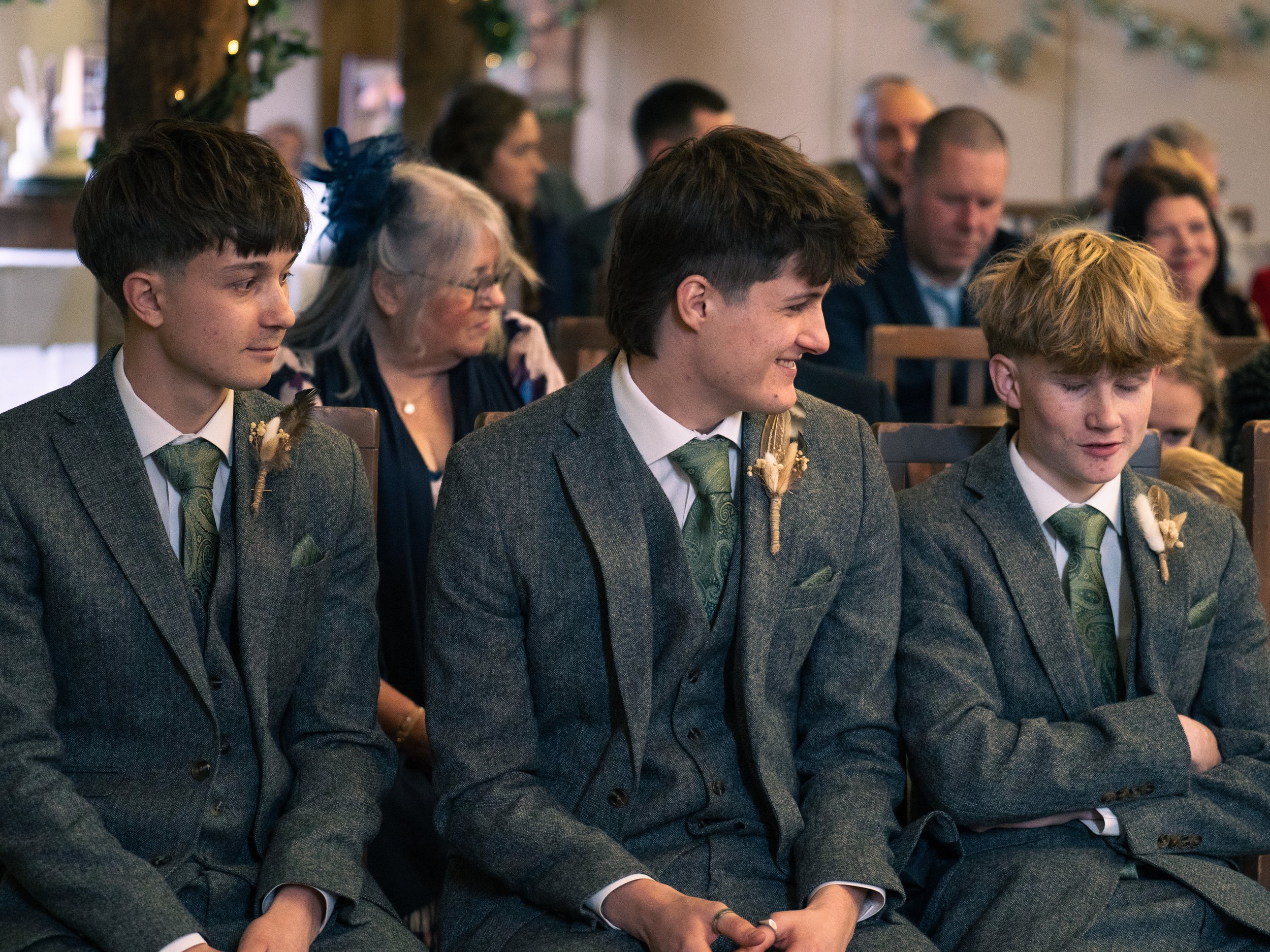 Three young boys in suits sitting in a row at a formal event, smiling and engaging with each other, with an older woman and more people seated behind them.