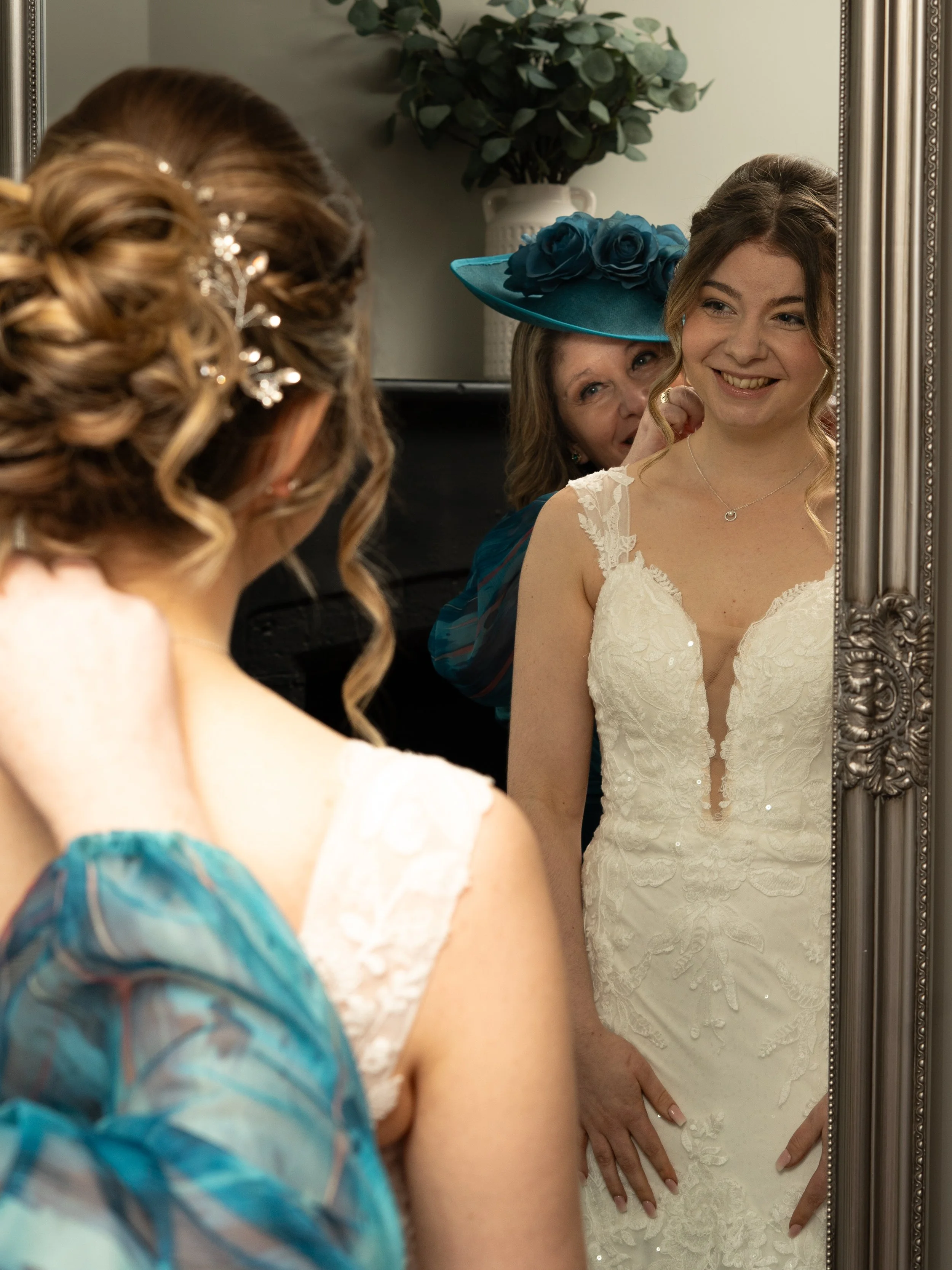 A woman in a white lace wedding dress is smiling in front of a mirror, with a woman wearing a blue hat with roses helping her adjust her hair.