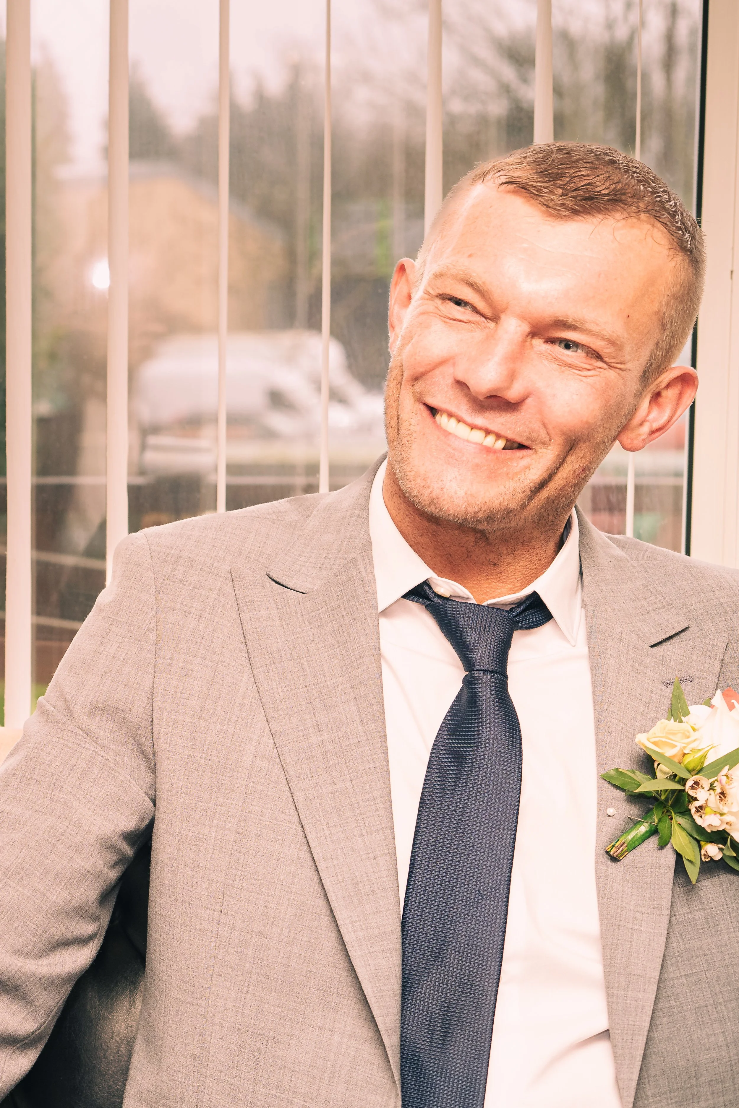 A smiling man in a gray suit with a white shirt and dark blue tie, wearing a boutonniere, sitting in front of a window with blinds.