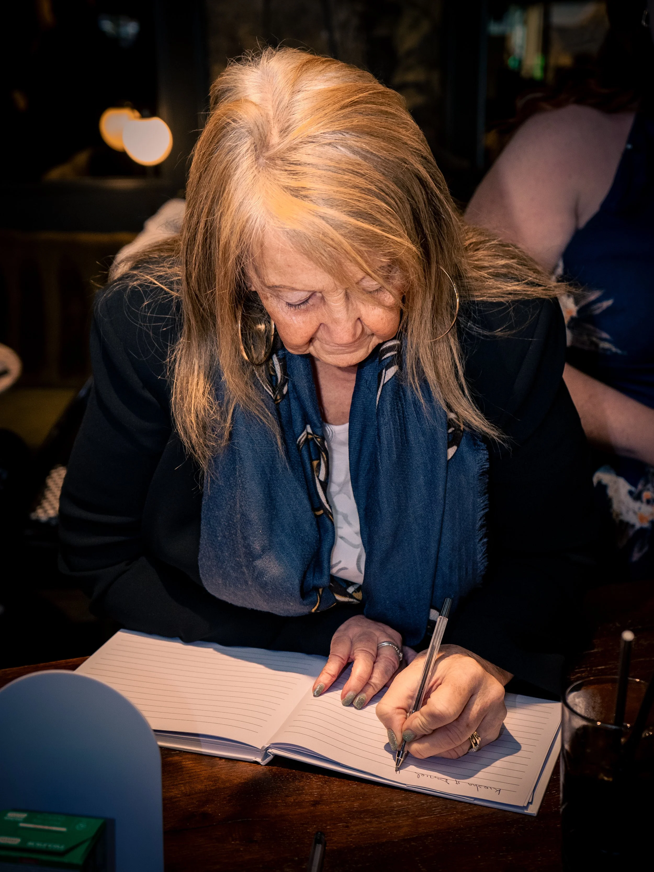 A woman with red hair and a blue scarf is writing in a journal at a table in a dimly lit setting.