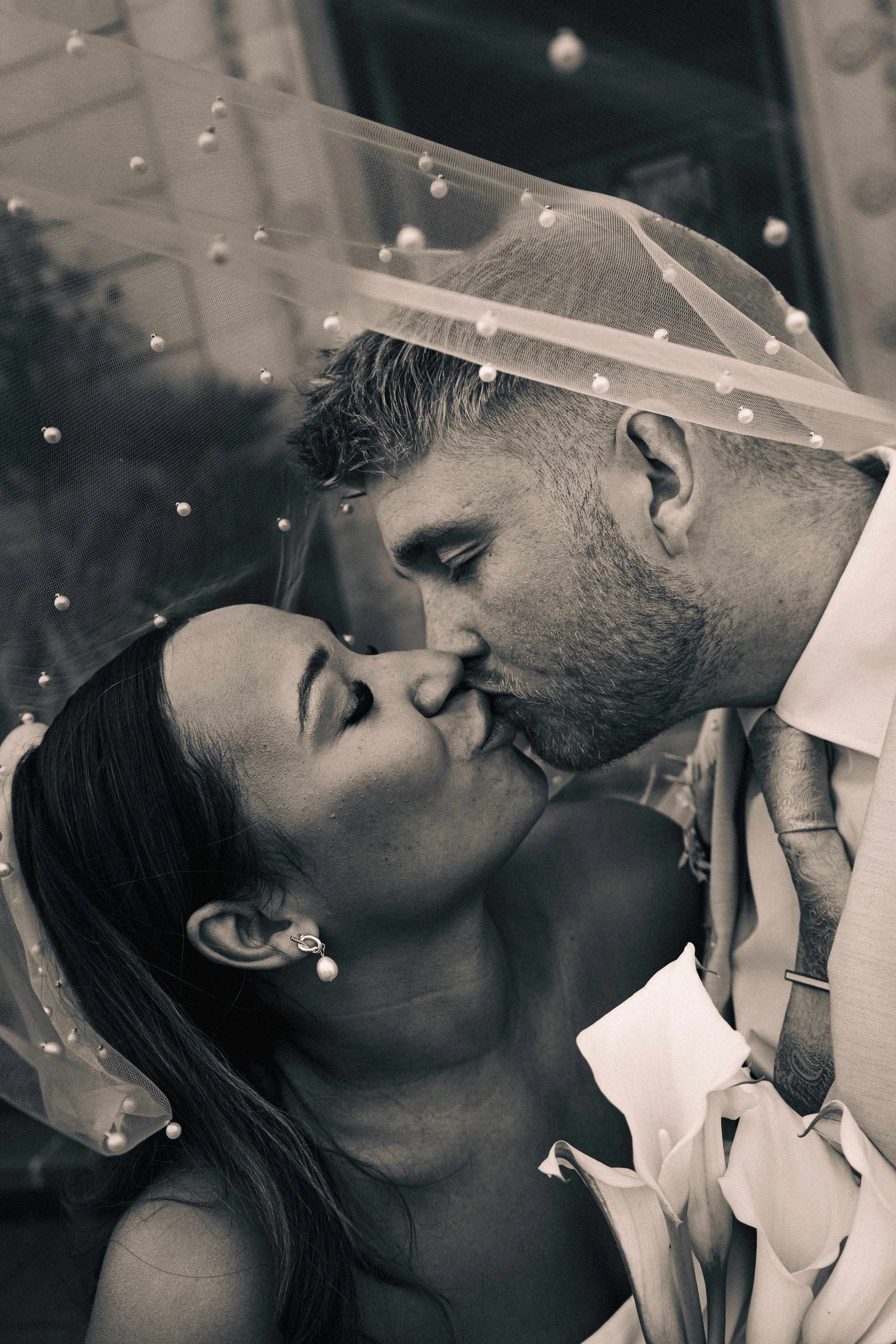 A black and white photo of a bride and groom kissing under a sheer veil, with the bride wearing earrings and holding calla lilies.