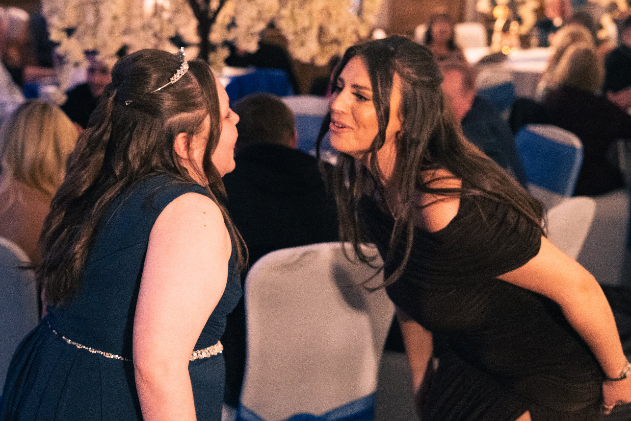 Two women are engaged in a close conversation at a formal event or wedding reception, with other guests seated at tables in the background.