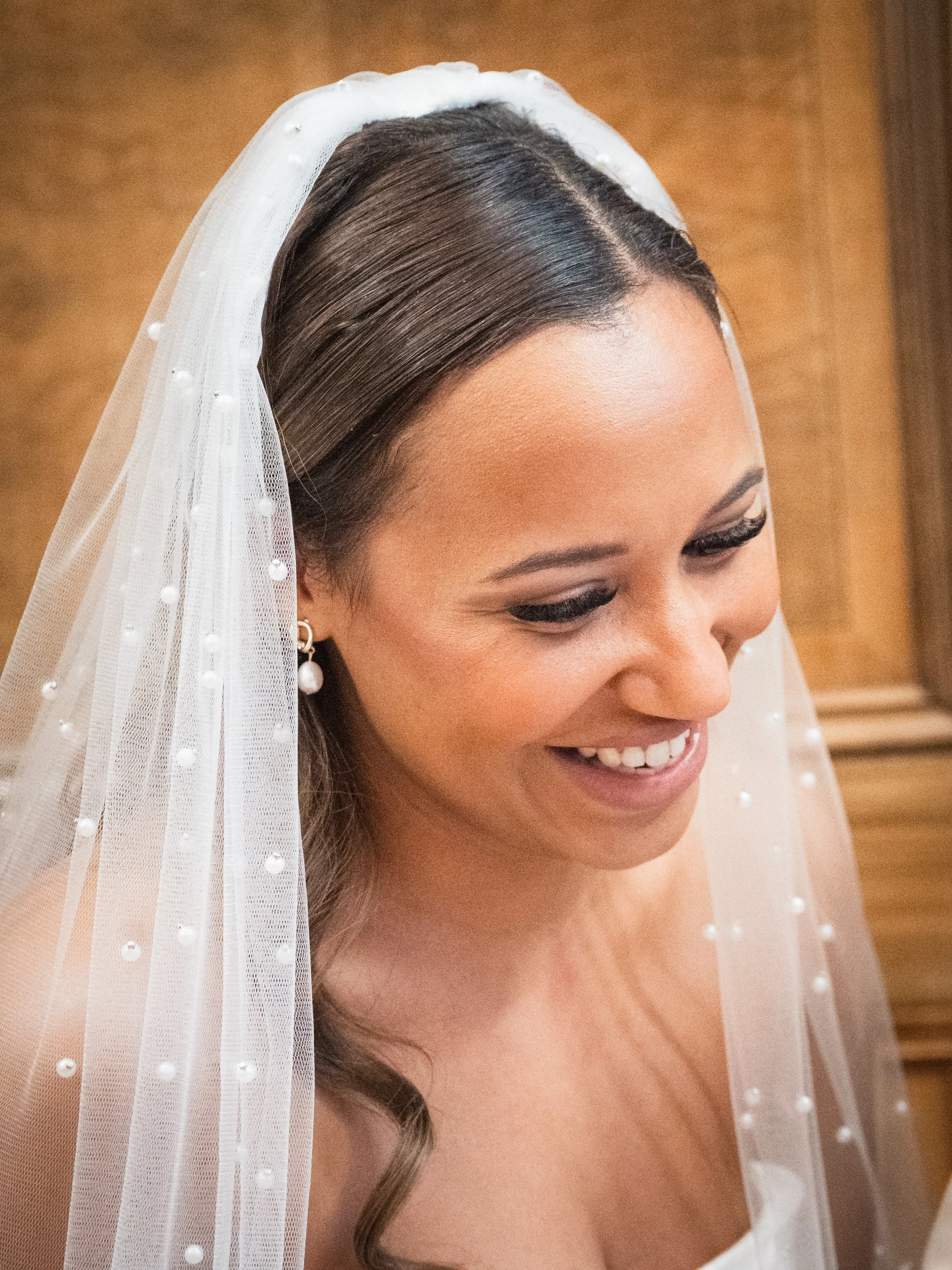 Close-up of a smiling bride with dark hair, wearing pearl earrings and a sheer veil with pearl embellishments.