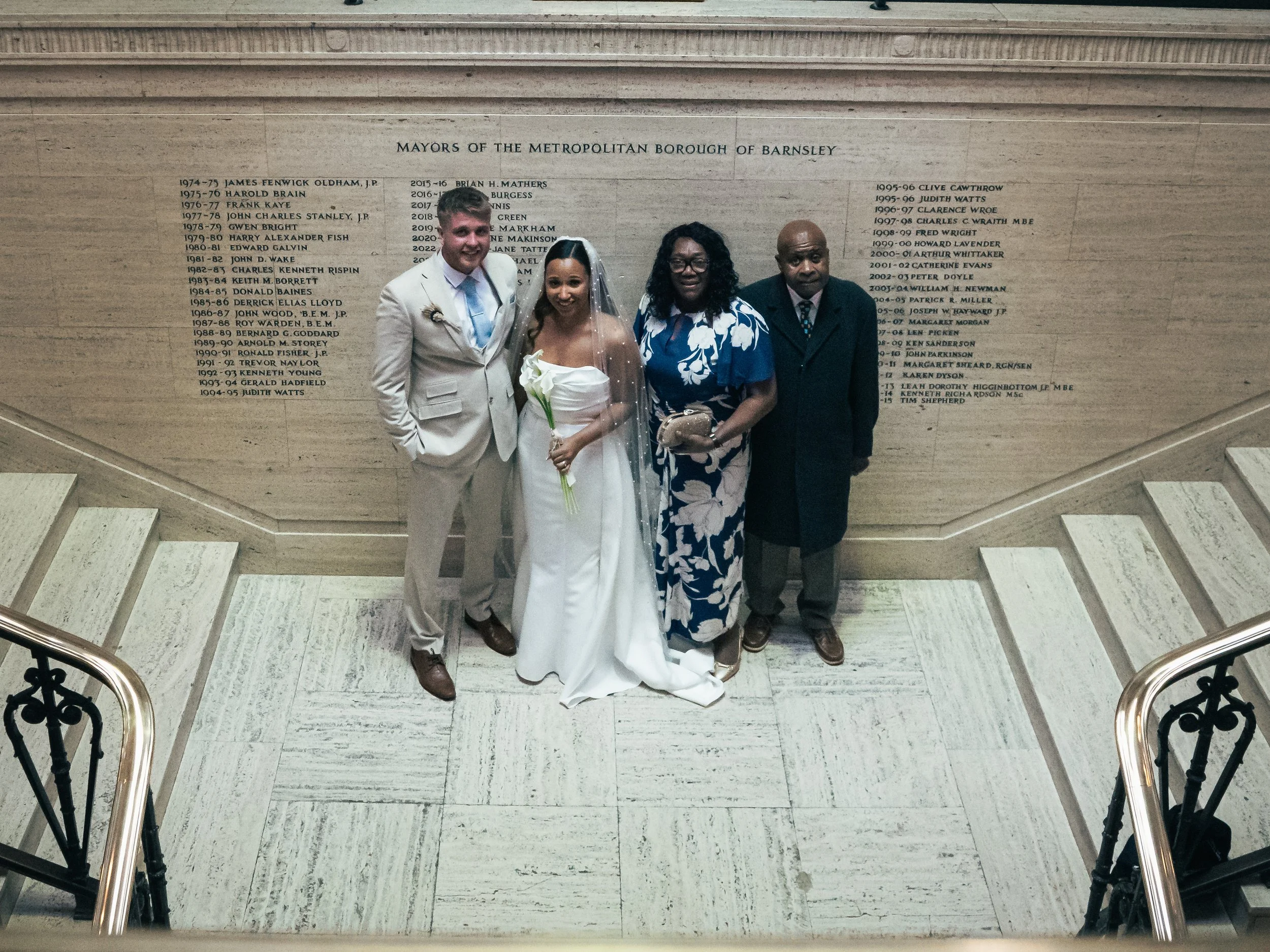 Group of five people standing on stairs in front of a tribute wall at Barnsley, including a bride in white dress and veil, five men, and a woman in a blue and white floral dress.