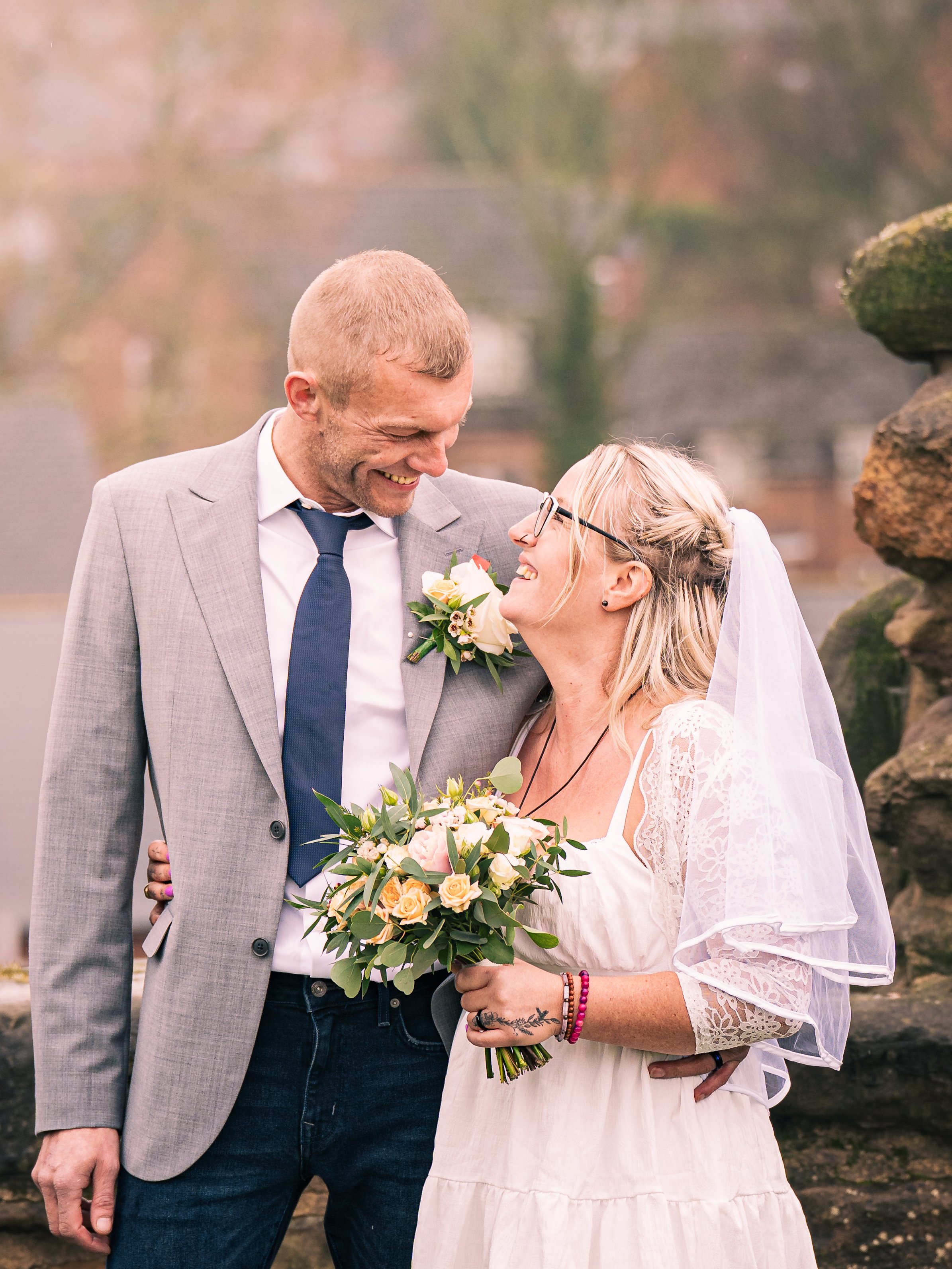 A newlywed couple shares a joyful moment outdoors, with the groom in a light grey suit and navy tie, and the bride in a white dress holding a bouquet, both smiling affectionately at each other.