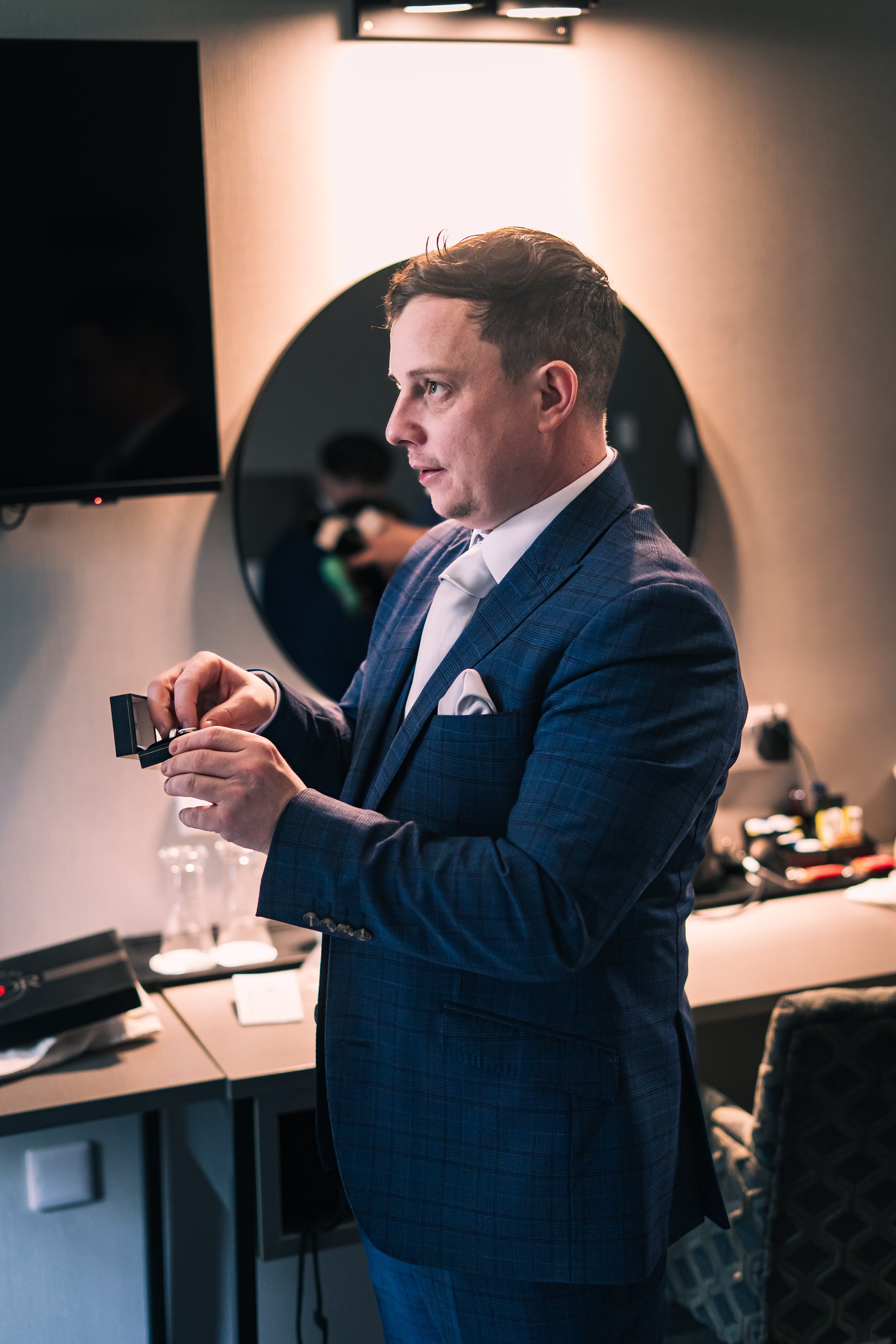 A man in a blue checkered suit adjusting a cufflink in a hotel room or dressing area with a large mirror and a TV on the wall.