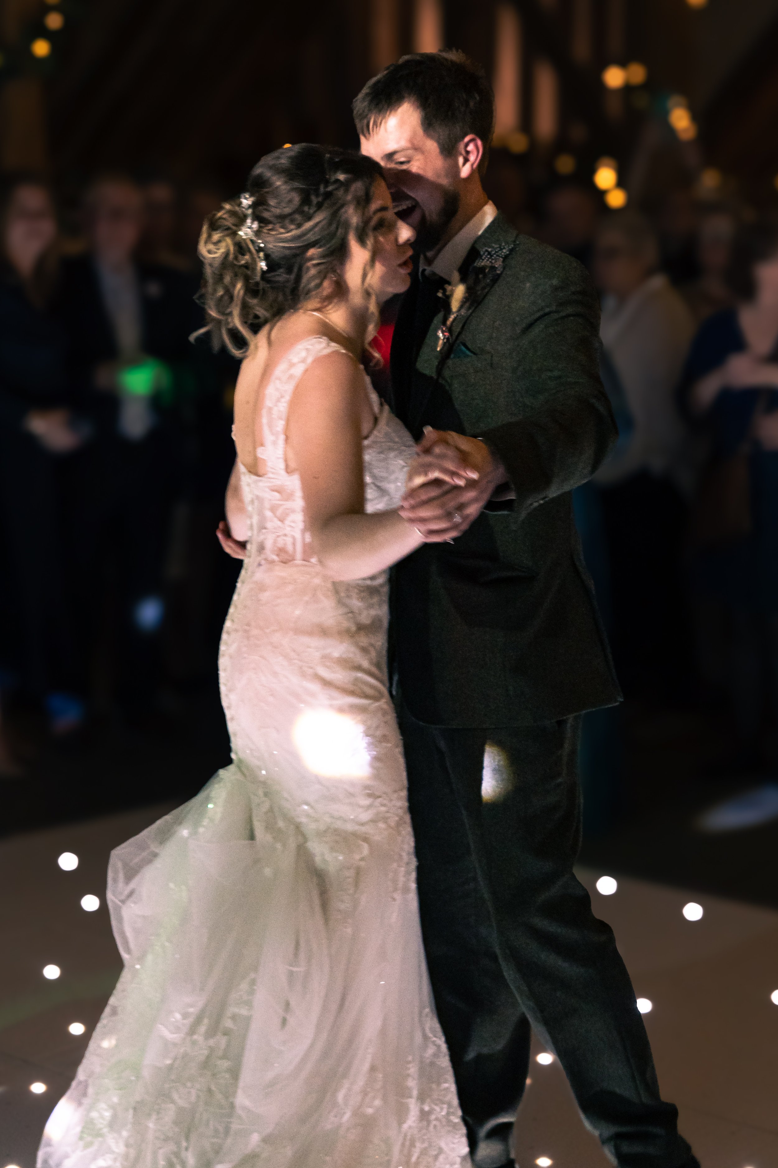 A bride and groom dancing closely at their wedding reception, surrounded by guests in a dimly lit venue.